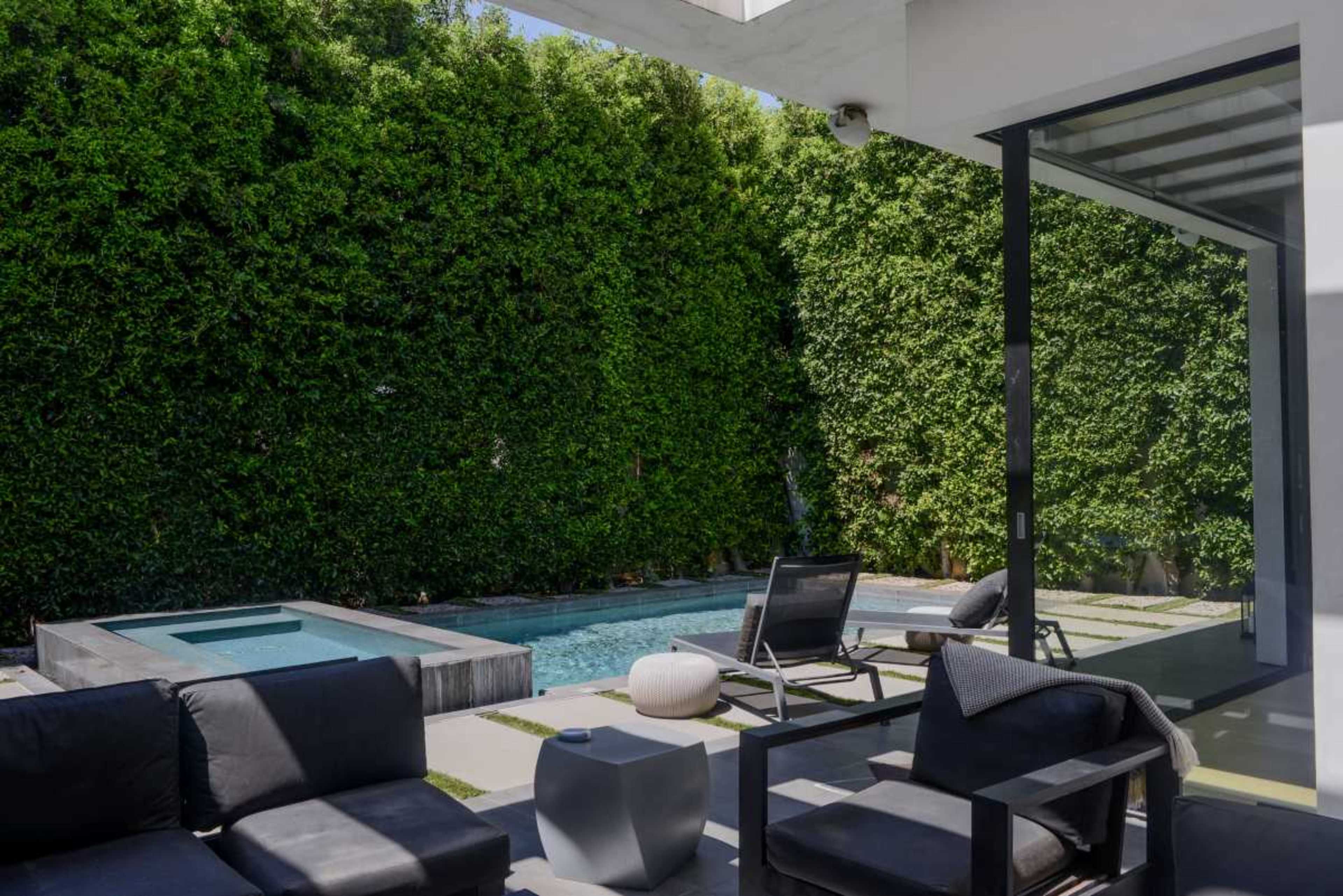 A small outdoor patio features black seating, a white side table, and a swimming pool surrounded by tall green hedges.