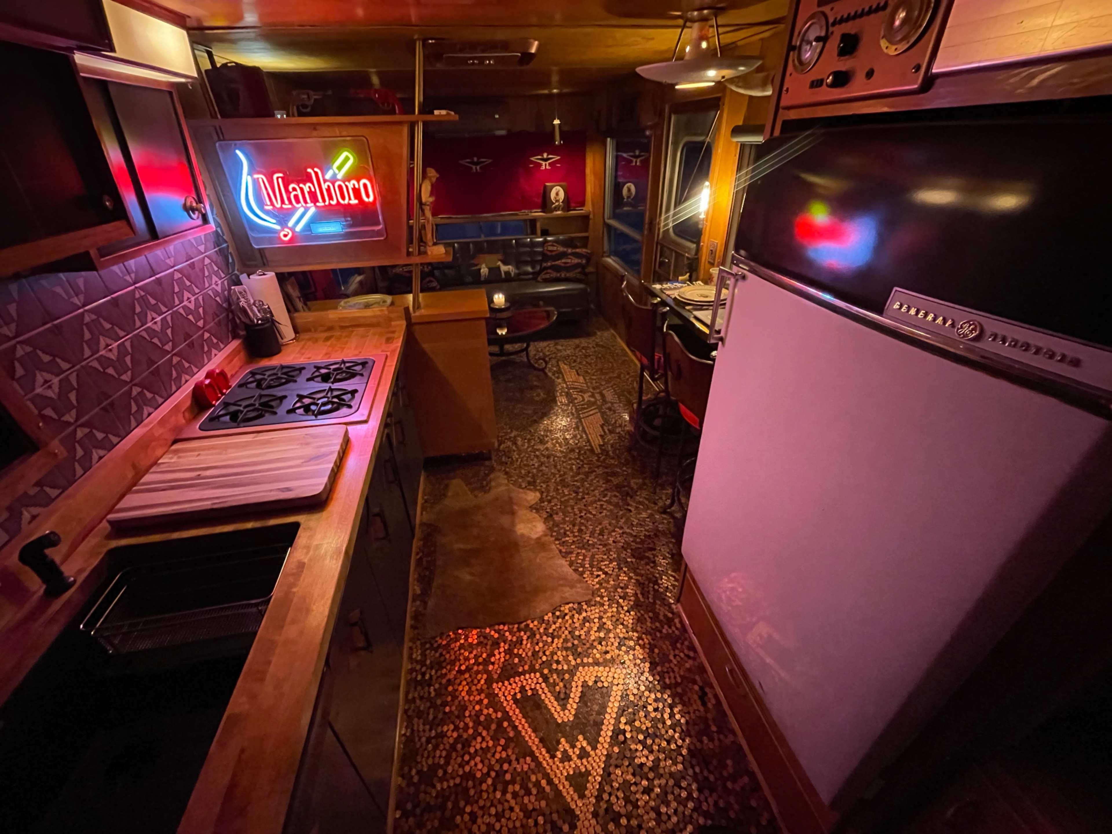 The image shows a well-lit kitchen area with wooden cabinets, a vintage fridge, a patterned floor, and a neon Marlboro sign.