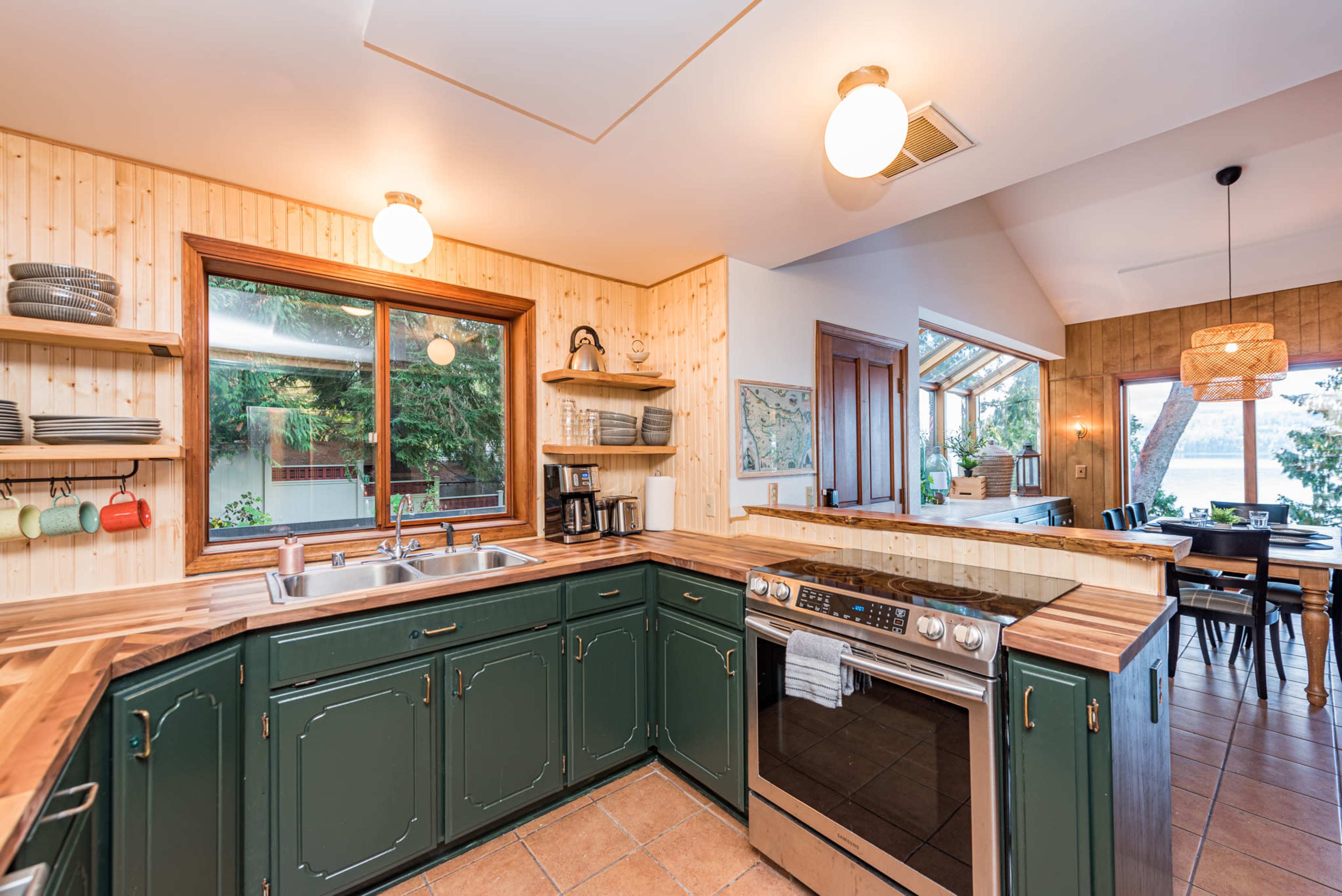 A kitchen with green cabinetry, a double sink, wooden countertops, and large windows overlooking an outdoor space.