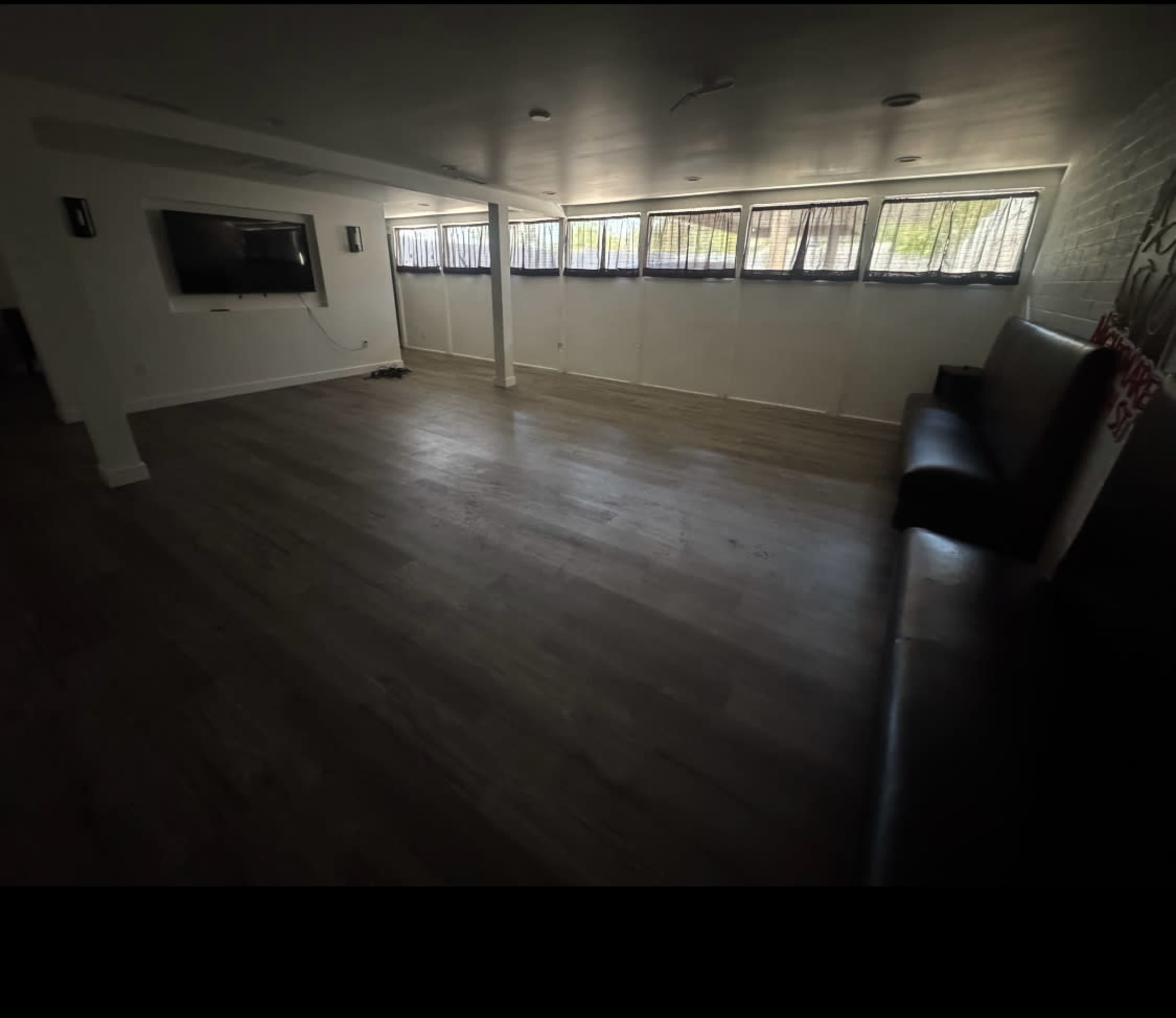 The image shows a spacious, empty room with light-colored walls, a flat-screen TV mounted on one wall, and several windows along another, allowing natural light to enter.