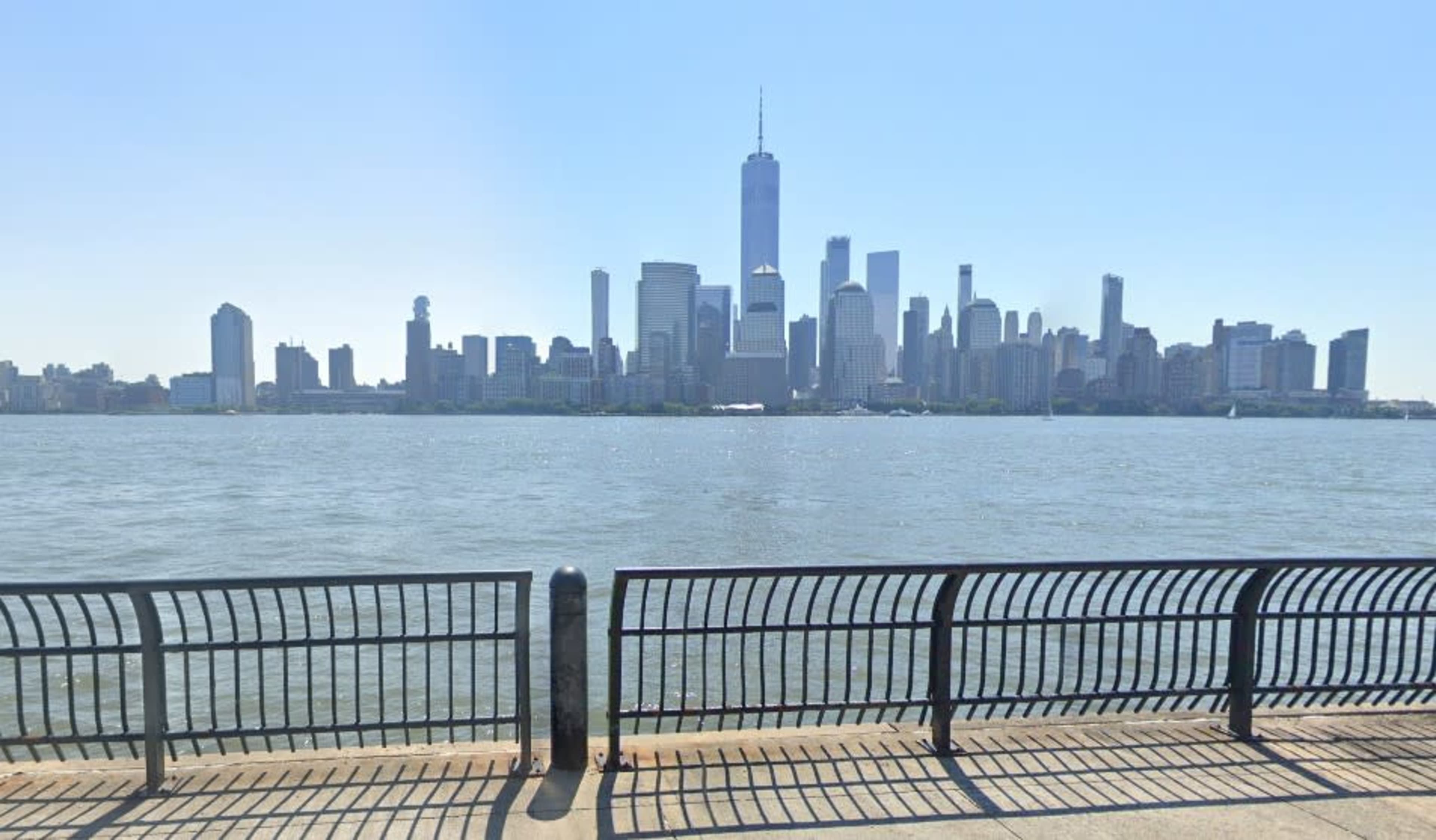 The image shows a view of a skyline featuring tall buildings, including One World Trade Center, across a river with a black railing in the foreground.