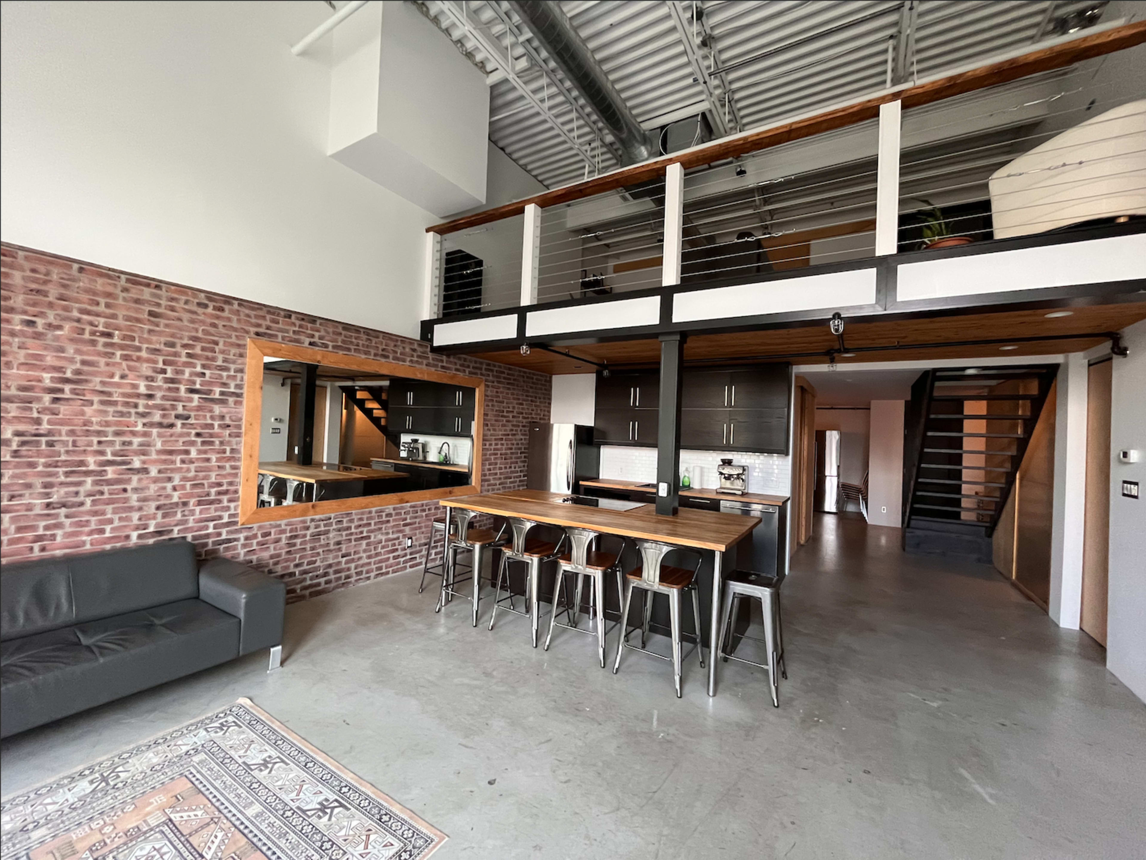A modern open-concept living space featuring exposed brick walls, a kitchen with bar seating, and a lofted area above.