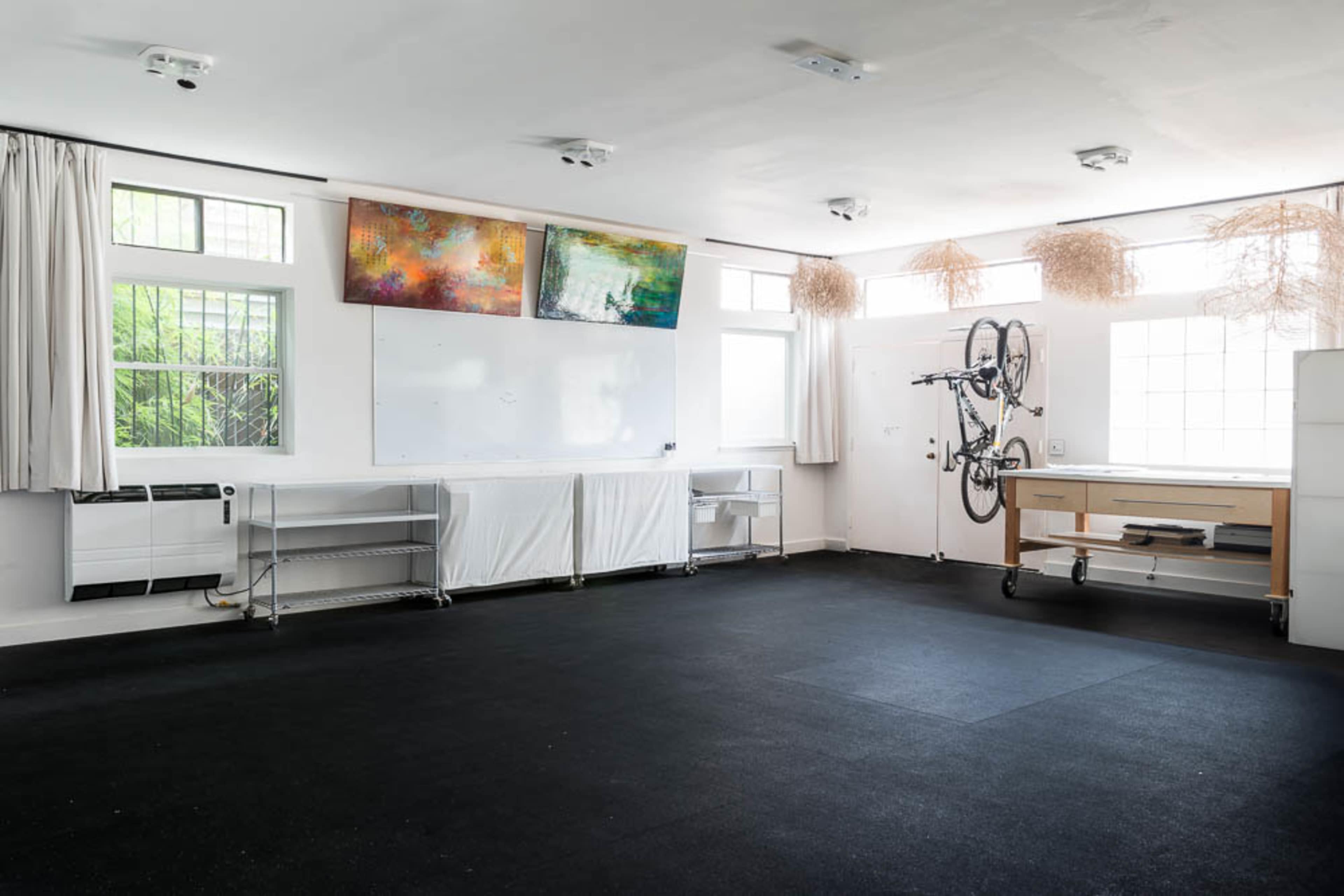 A spacious, empty room with white walls, large windows, two abstract paintings, and a bicycle mounted on the wall.