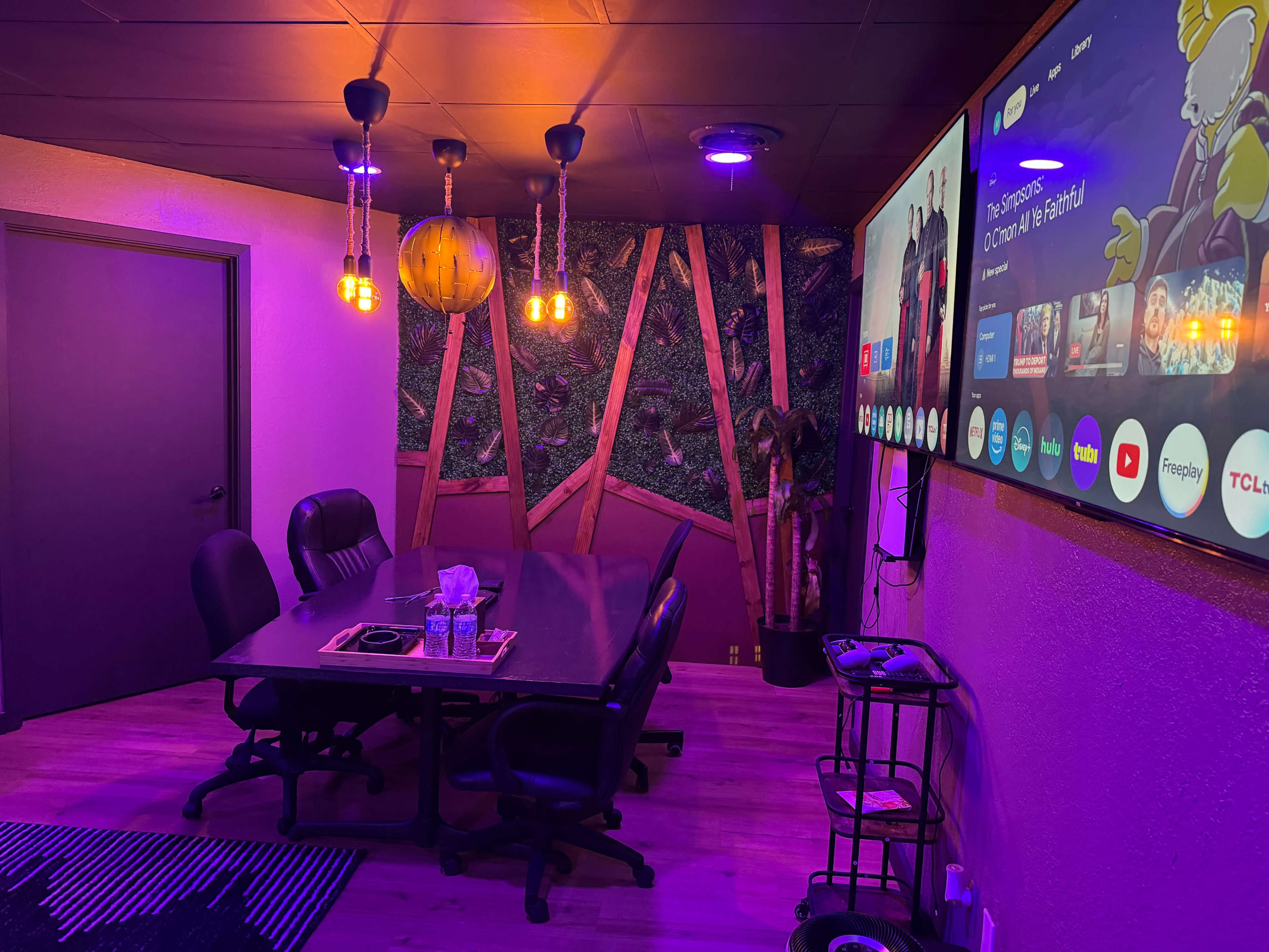 The image shows a modern meeting room with a large table, black chairs, and a wall-mounted screen, illuminated by colorful lights and decorative wall features.