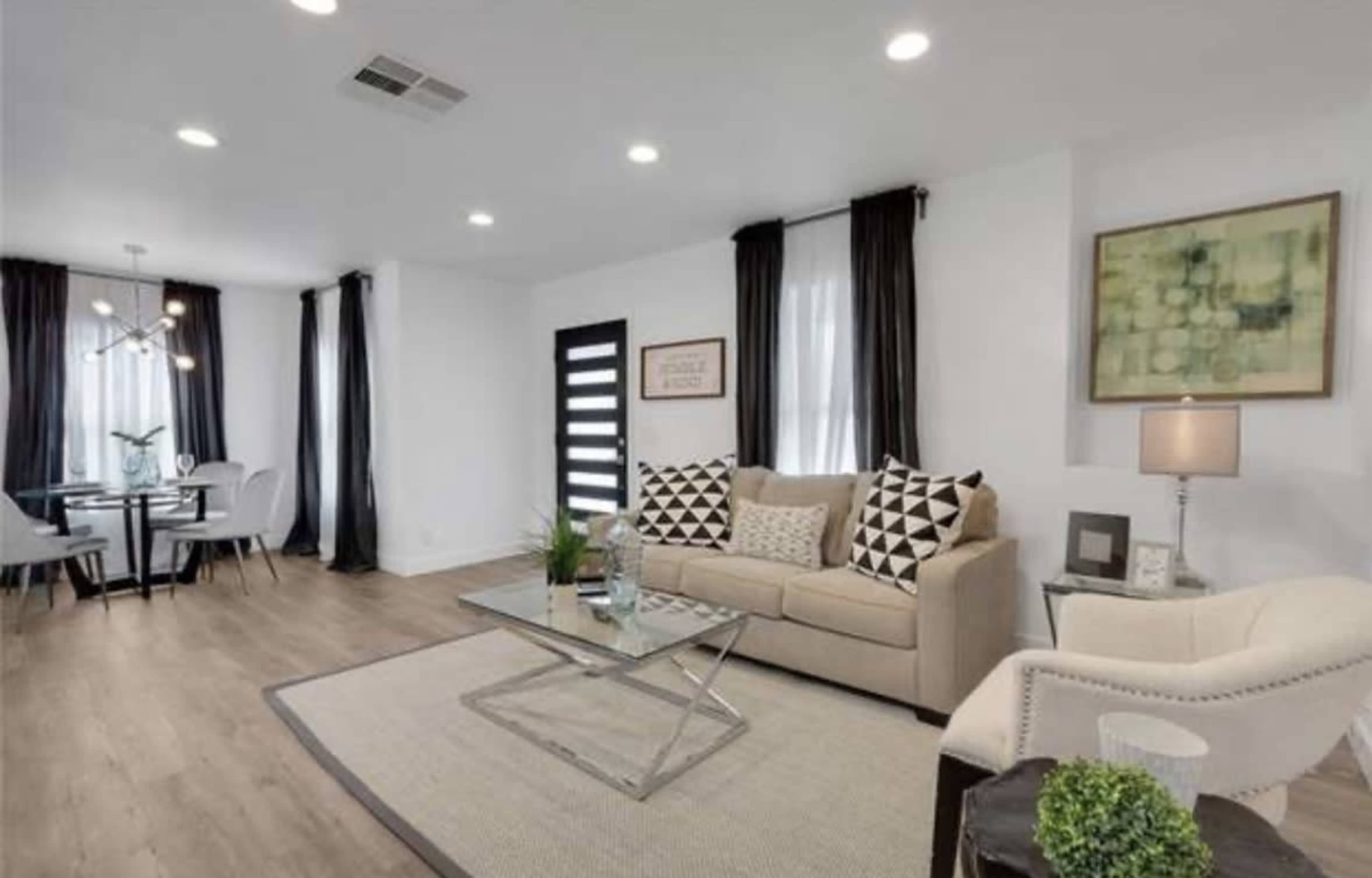 A modern living room with a beige sofa, black and white patterned pillows, a glass coffee table, and a dining area with a table and chairs in the background.