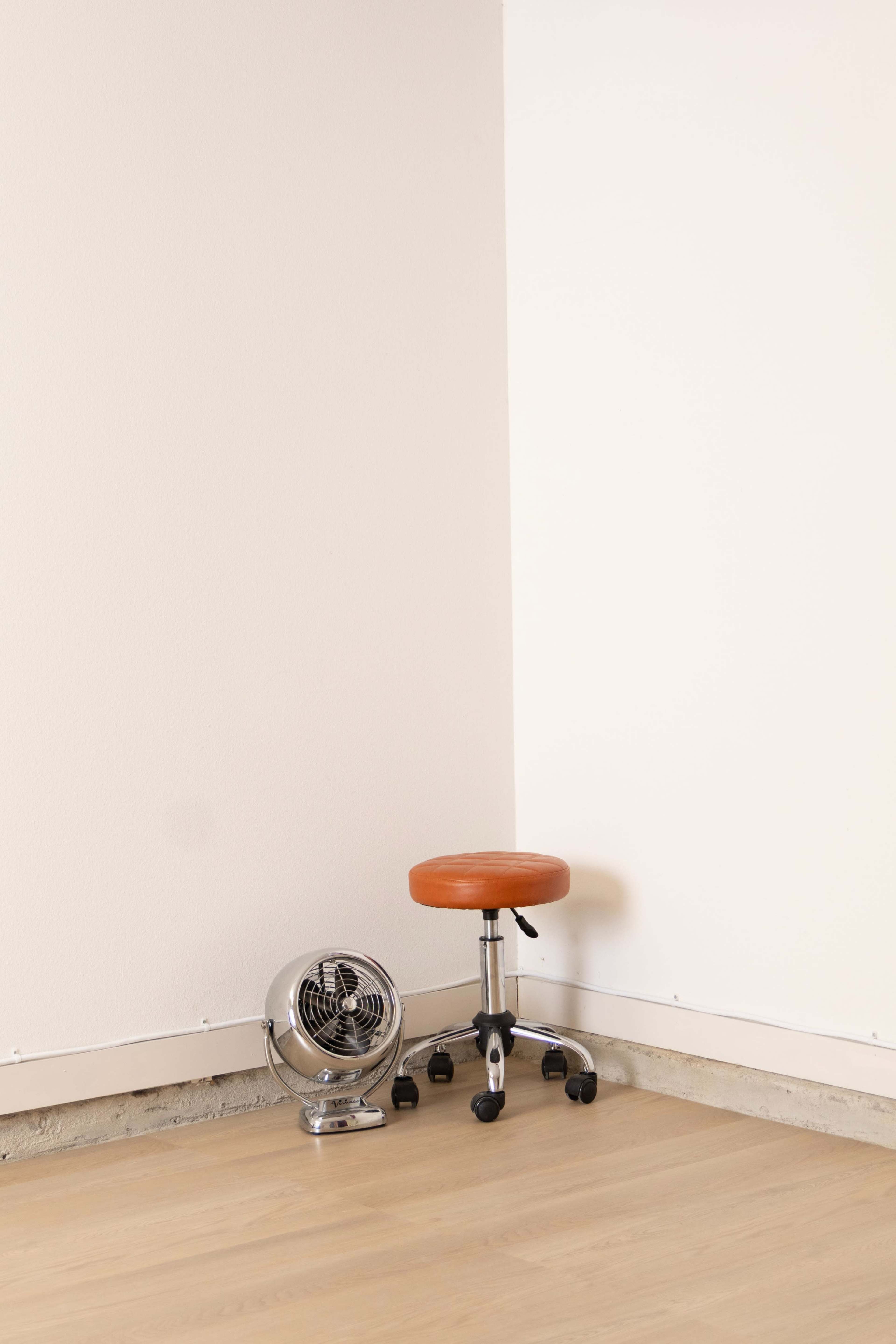 A wooden stool with an orange seat and a small metal fan are positioned in the corner of a room with light-colored walls and a wooden floor.