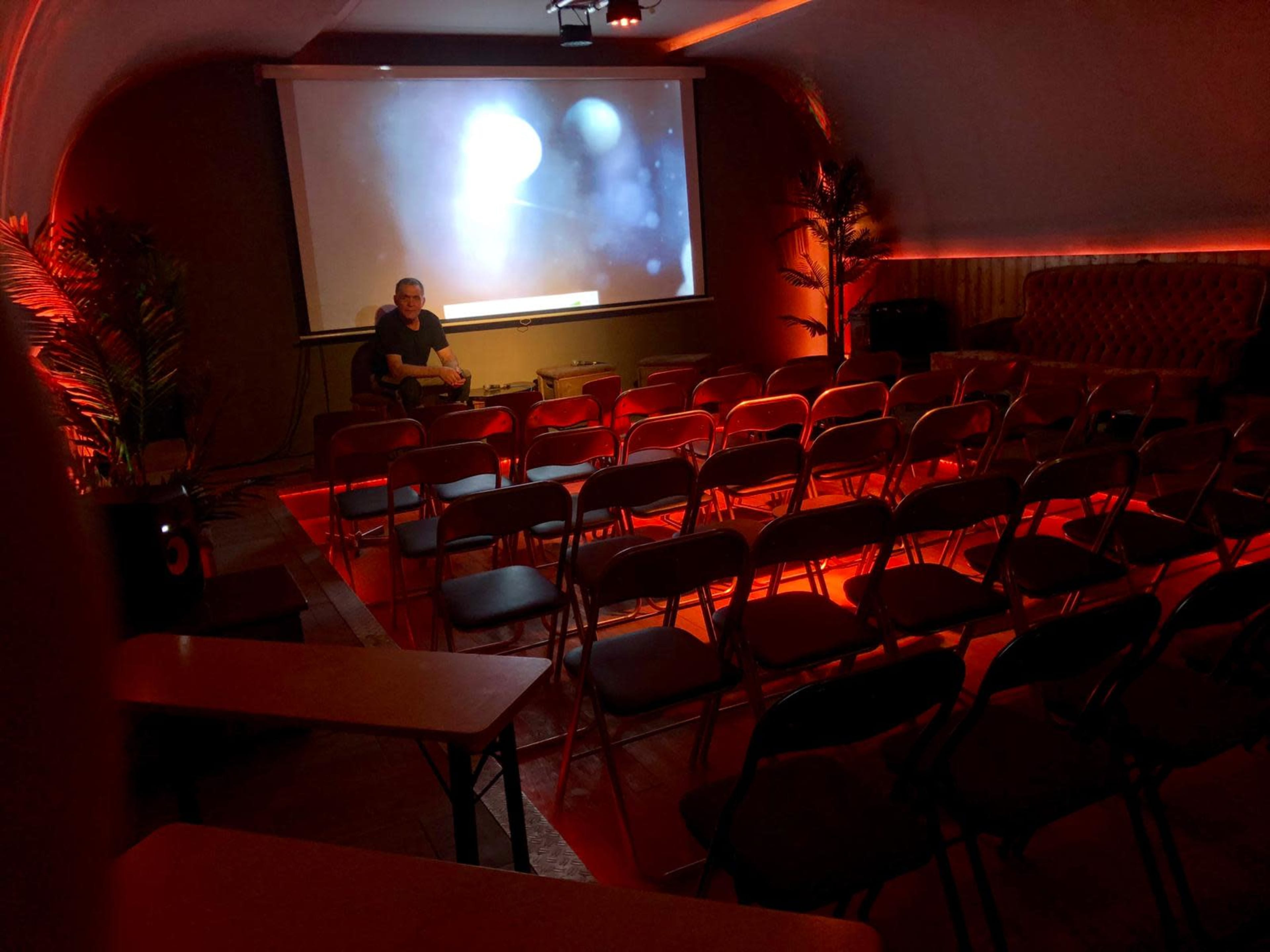 A dimly lit room with rows of empty folding chairs facing a large screen, where a person sits in front of the display.