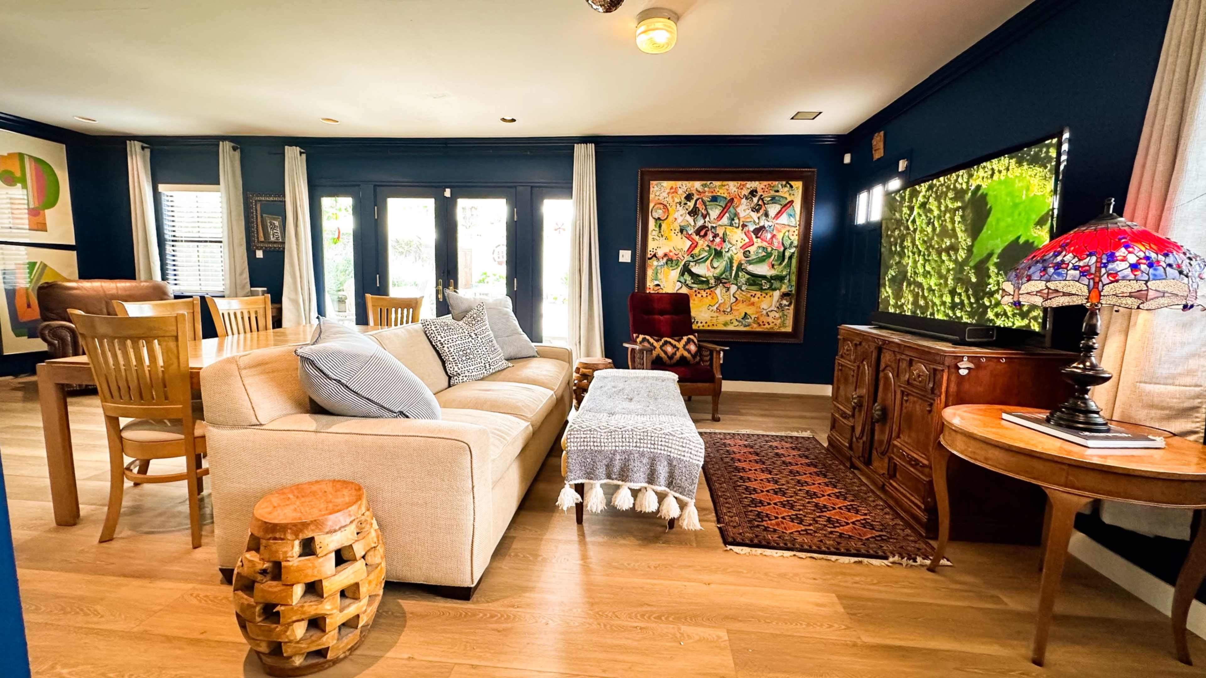 The living room features a beige sofa with blue pillows, a wooden dining table, a decorative rug, and a large framed artwork on the wall, alongside a television and a lamp.