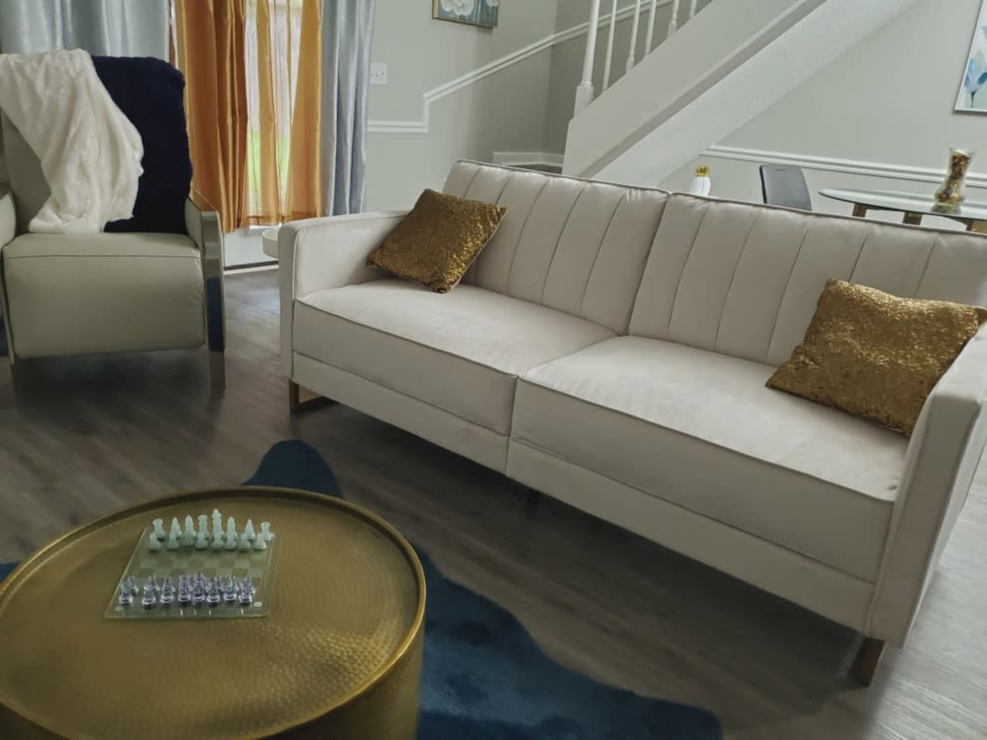 A modern living room featuring a light-colored sofa with gold-accented pillows, a round coffee table with a chess set, and curtains in shades of grey and orange.