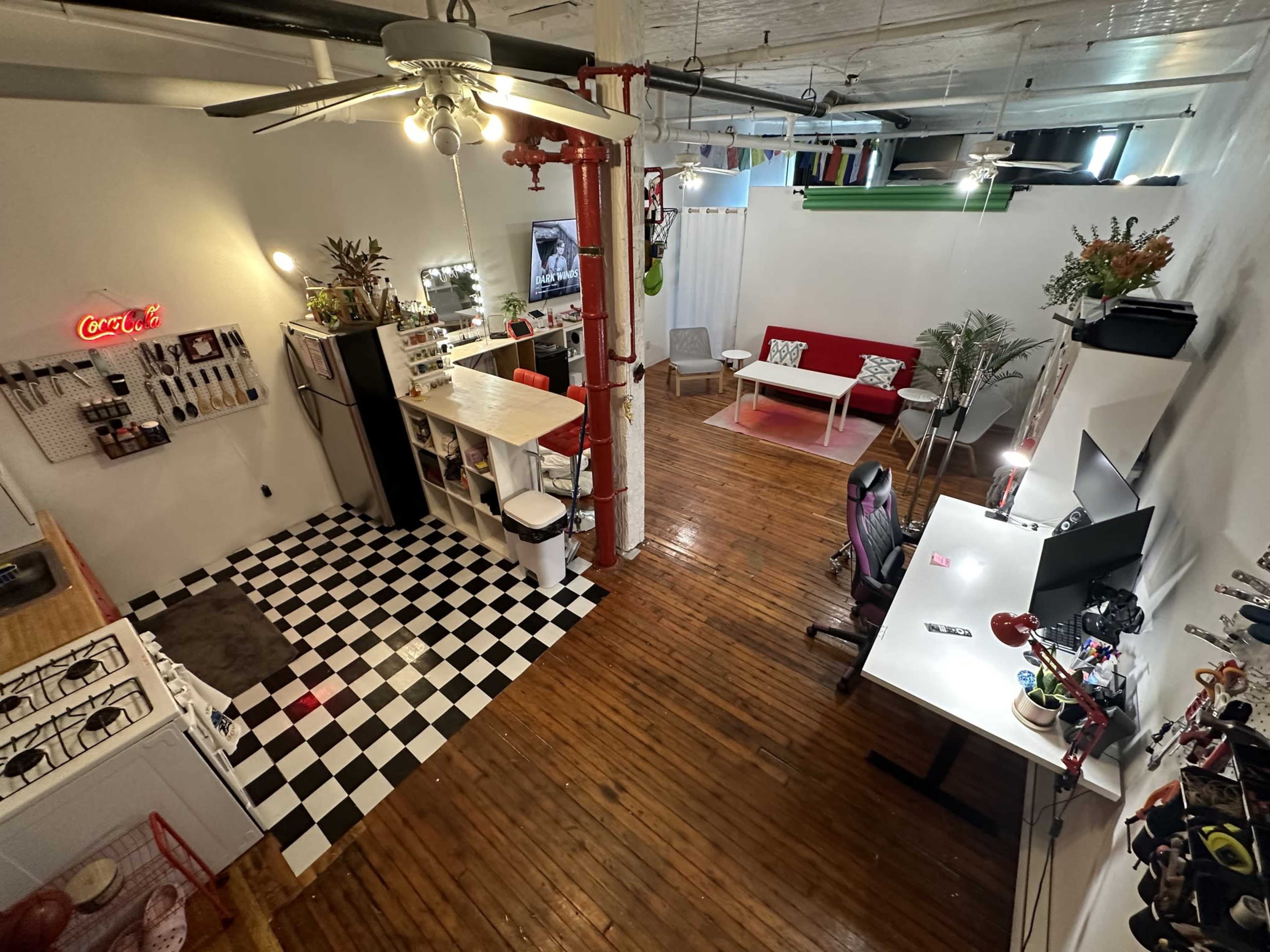 The image shows a spacious loft-style room featuring a kitchenette with black and white checkered flooring, a red sofa, a desk with a computer, and various decorative plants.
