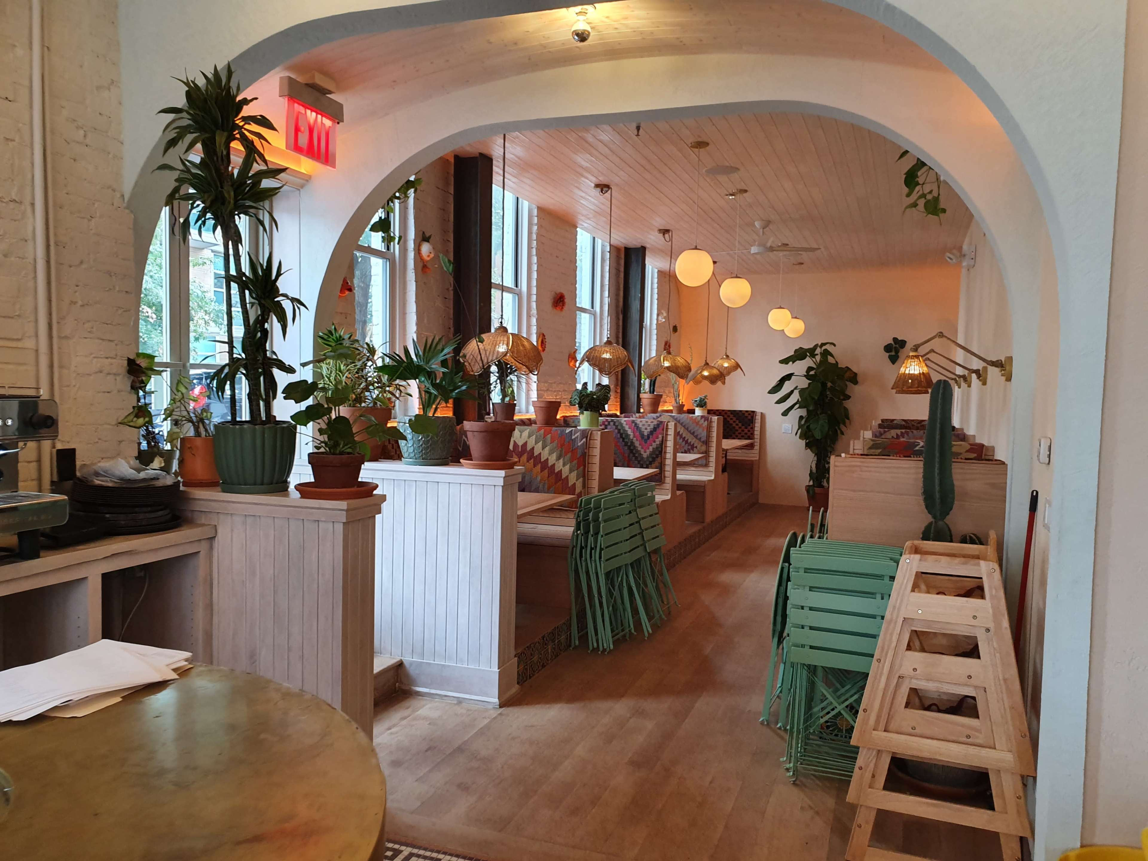 A brightly lit café interior featuring arched doorways, wooden ceiling, potted plants, and a mix of seating arrangements with decorative lighting.