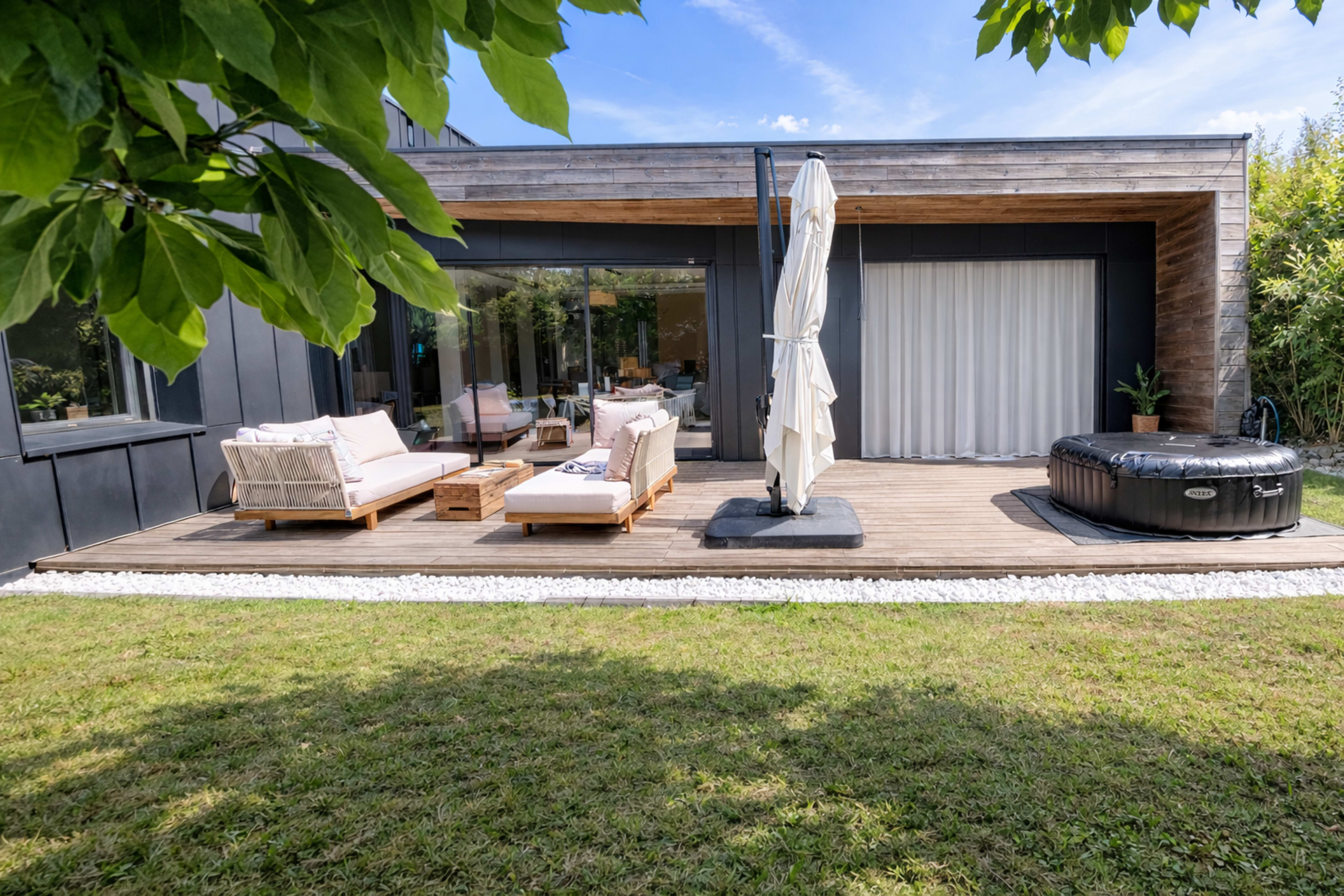 A modern house features a wooden deck with outdoor seating, an umbrella, and a black hot tub, surrounded by a grassy lawn.