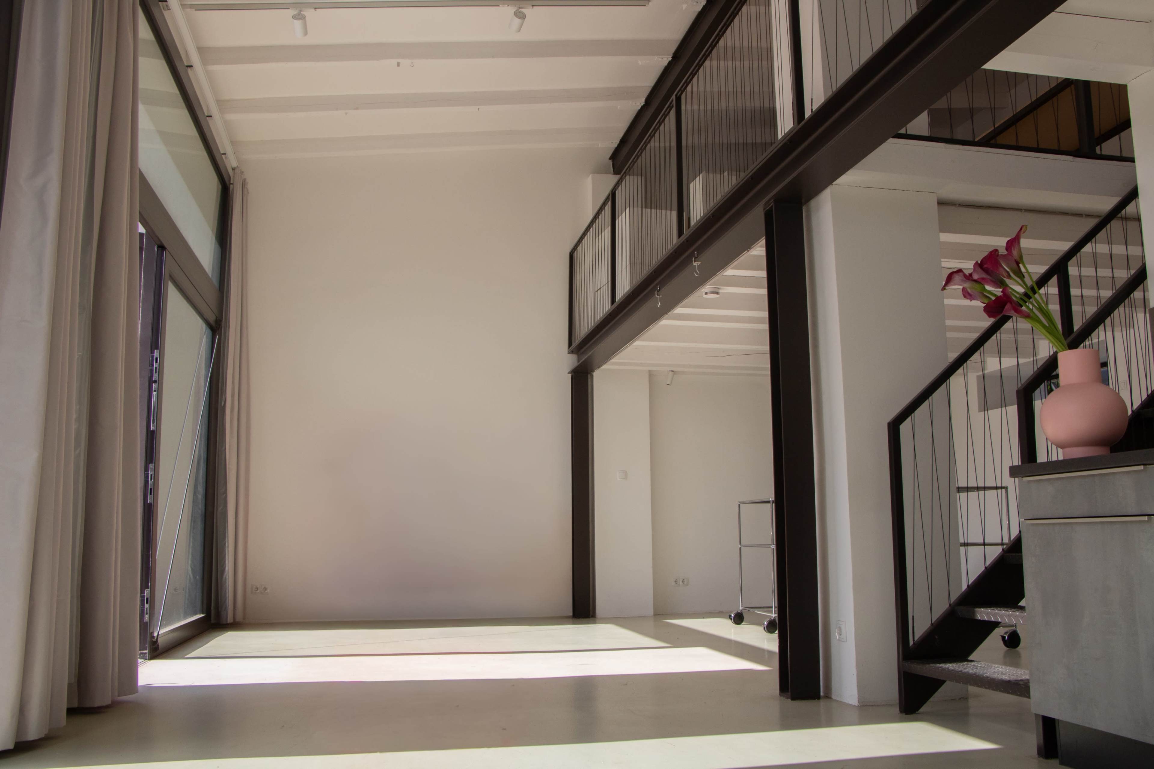 The image shows a spacious modern interior with large windows, a staircase, and a minimalistic decor featuring a vase with flowers on a table.