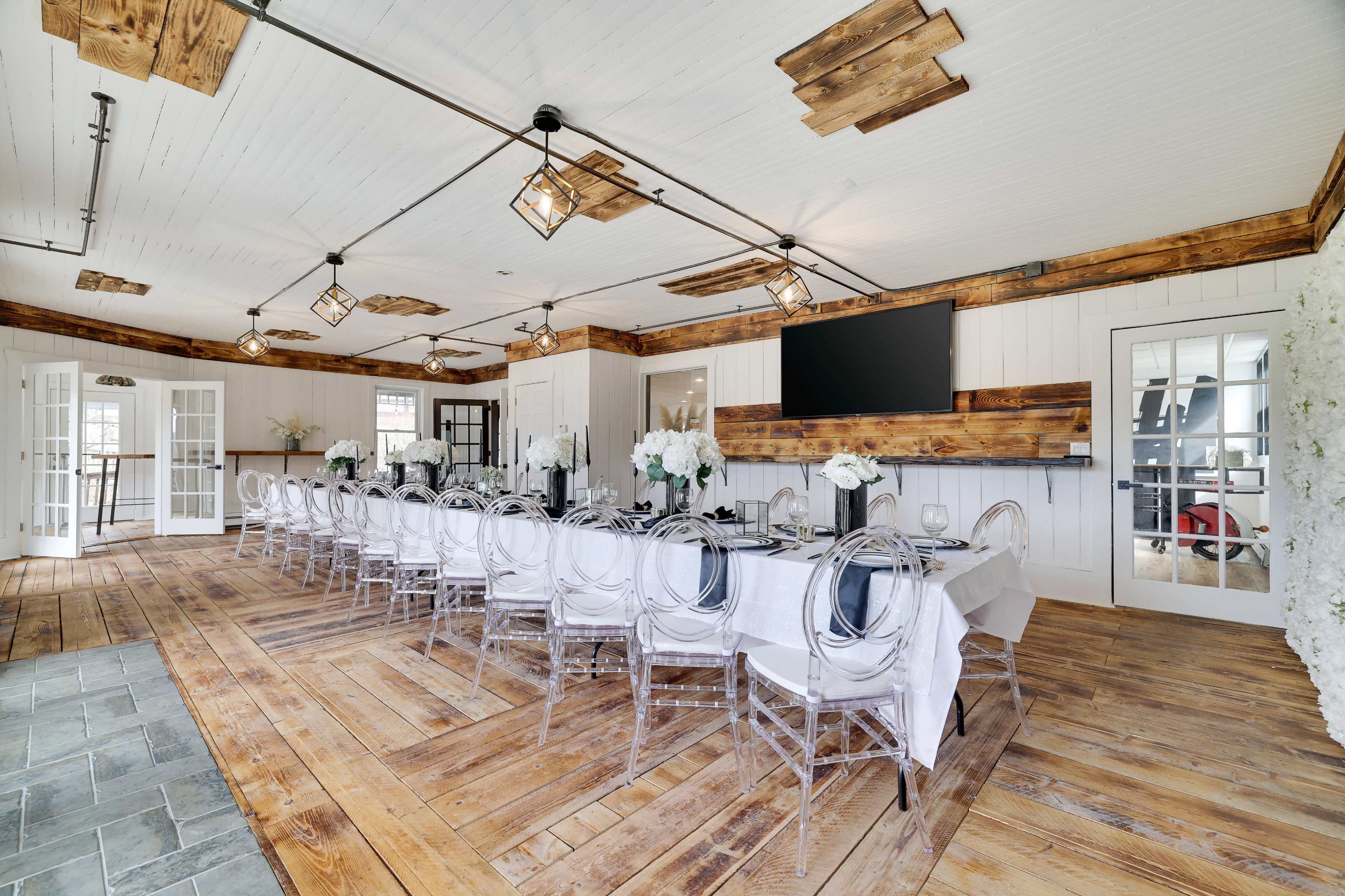 A long dining table is set with white tablecloths and clear chairs in a room with wooden beams, a large television, and white floral arrangements.