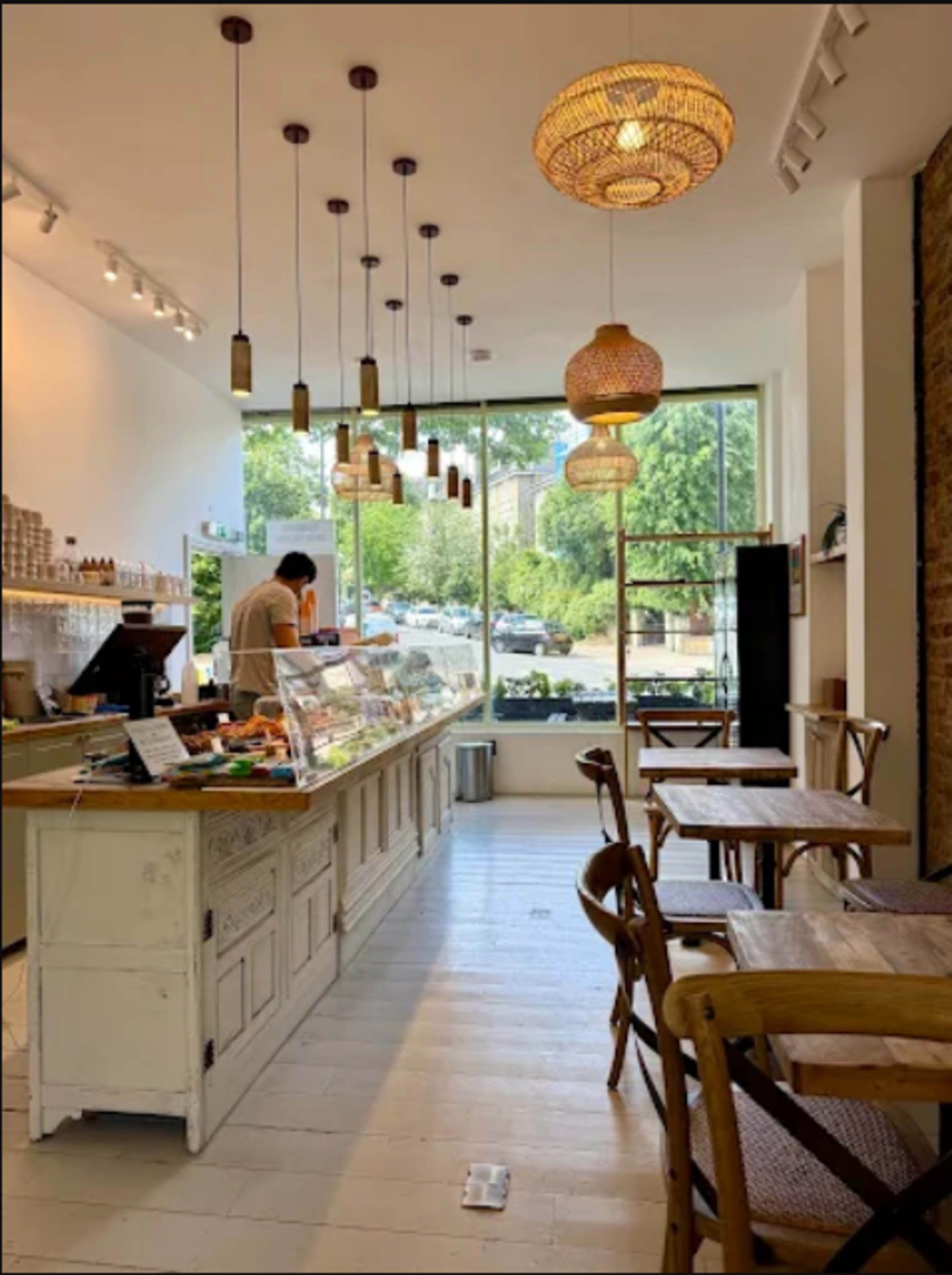 The image shows a bright café interior with a counter displaying desserts and several wooden tables arranged for seating.
