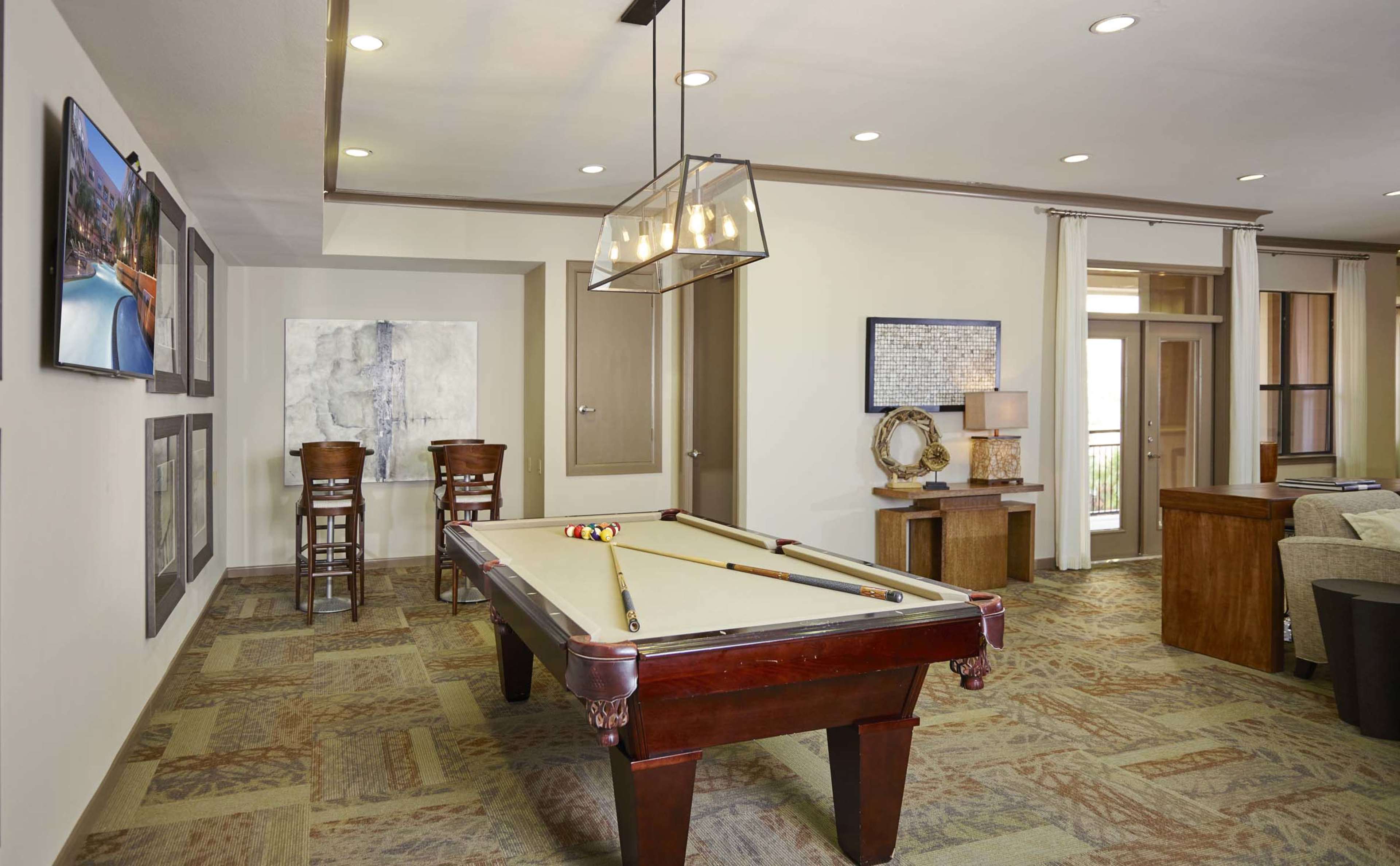A game room with a pool table, two bar stools, and a seating area, featuring modern decor and lighting.
