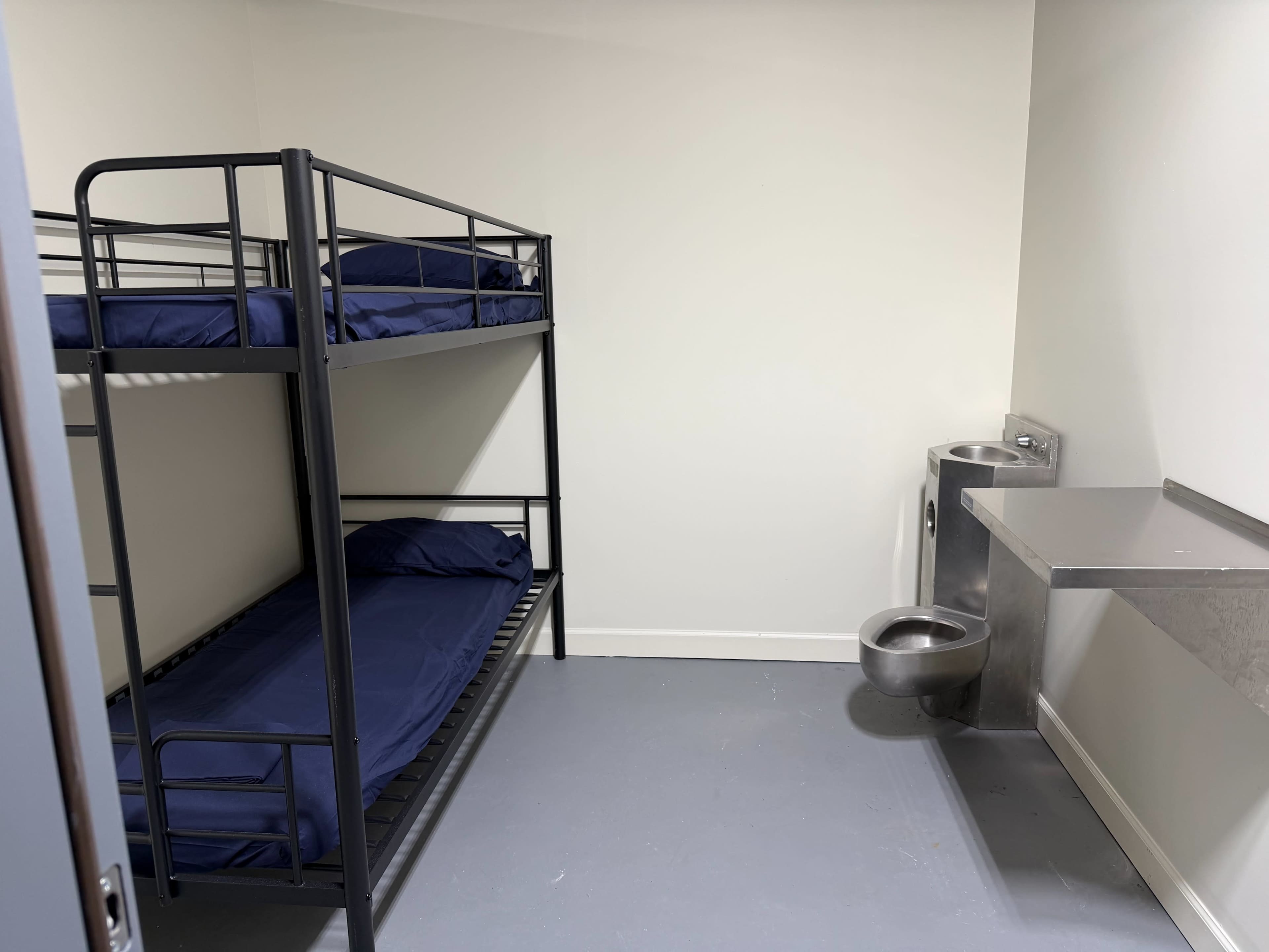 The image shows a small, bare room with a metal bunk bed, two mattresses, a wall-mounted sink, and a stainless steel table.