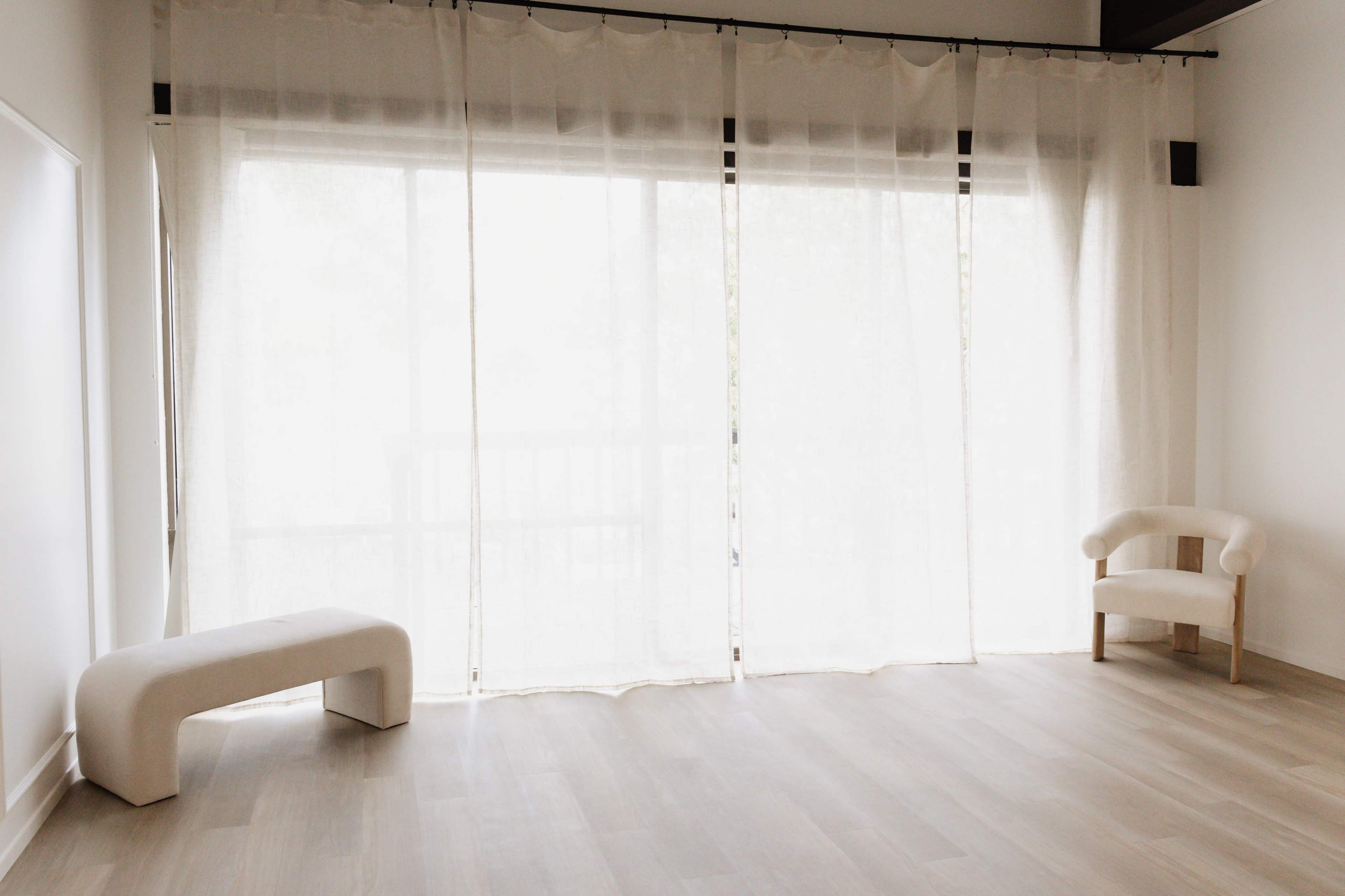 A light-filled room features sheer curtains covering a large window, a white bench, and a small, minimalist chair.