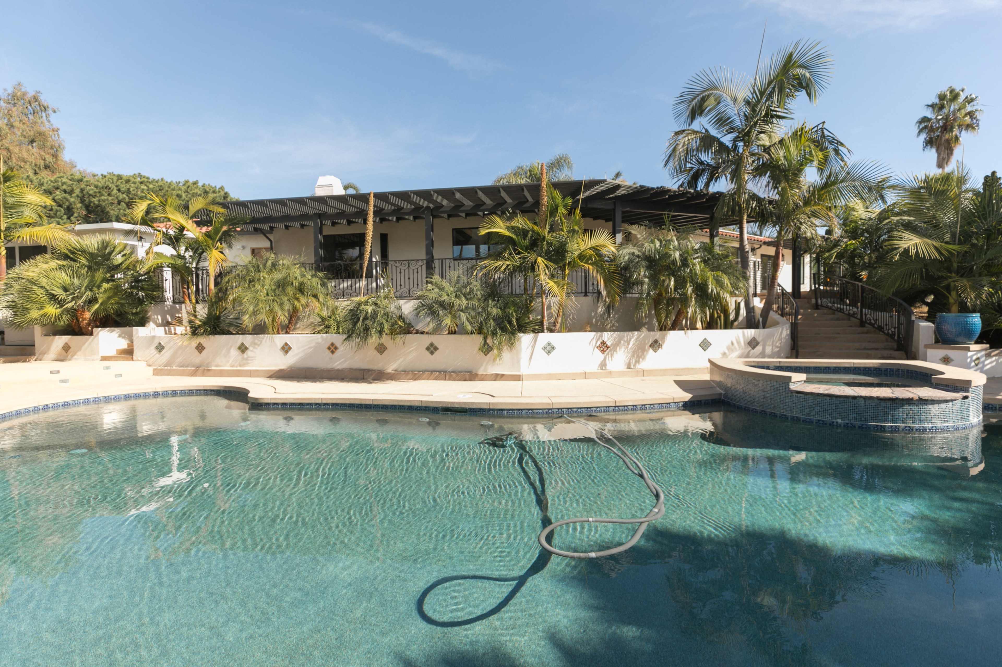 A landscaped backyard features a swimming pool with a jacuzzi, surrounded by palm trees and a house in the background.