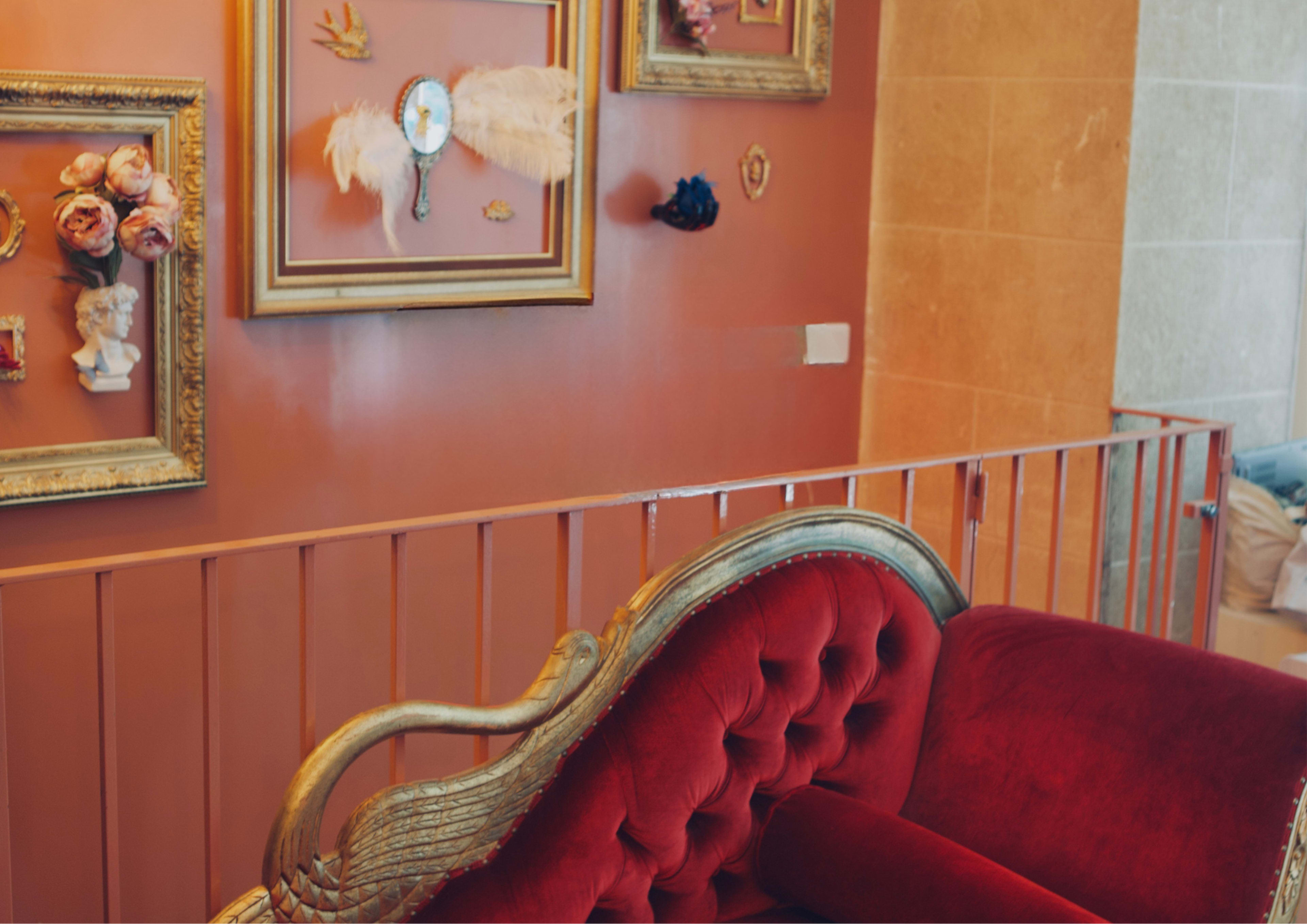 A red velvet couch with ornate gold detailing is positioned against a peach-colored wall adorned with framed artwork and decorative accents.