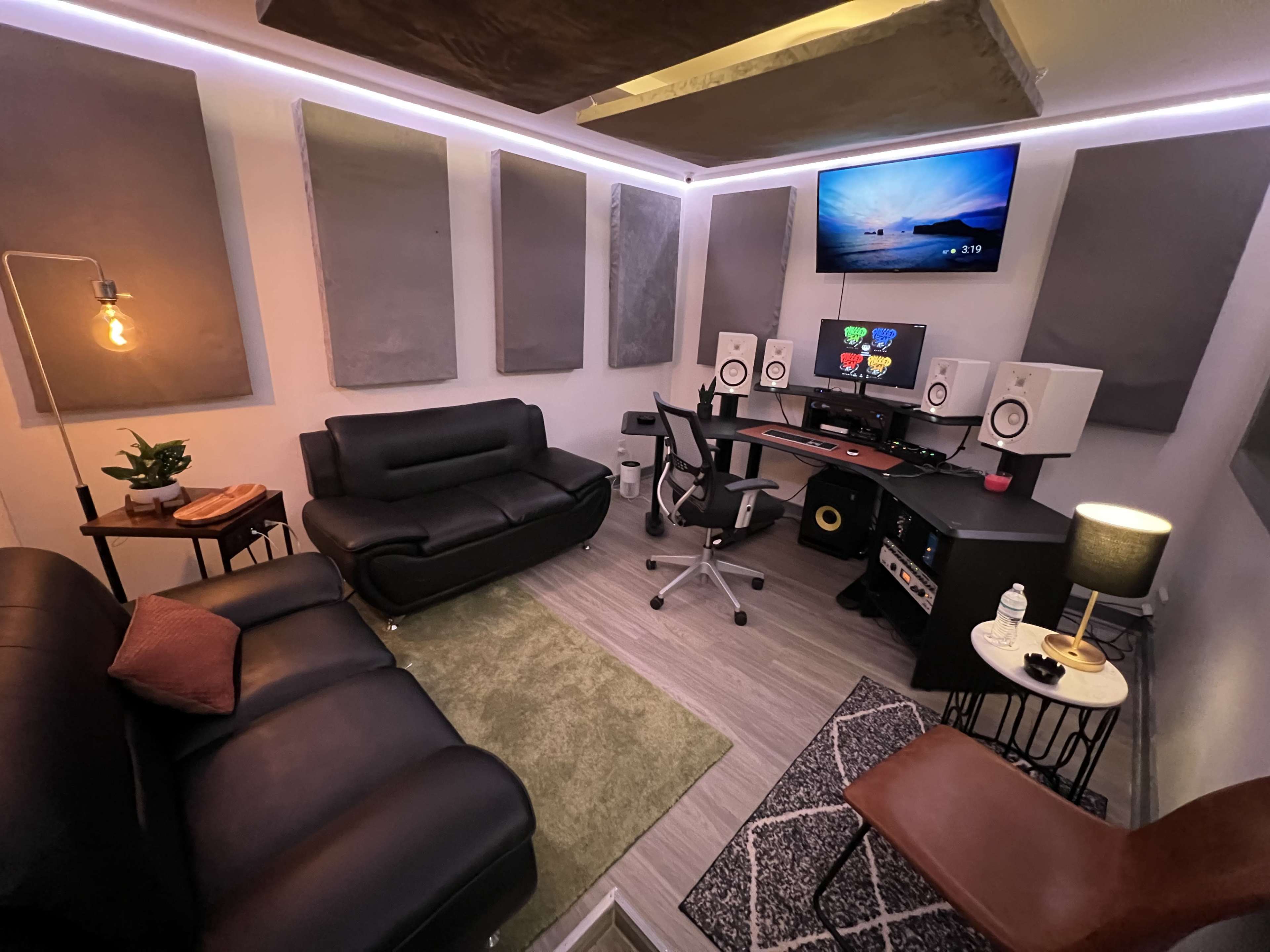 A compact music studio featuring a black sofa, a desk with speakers and a computer monitor, soundproofing panels on the walls, and various seating arrangements.