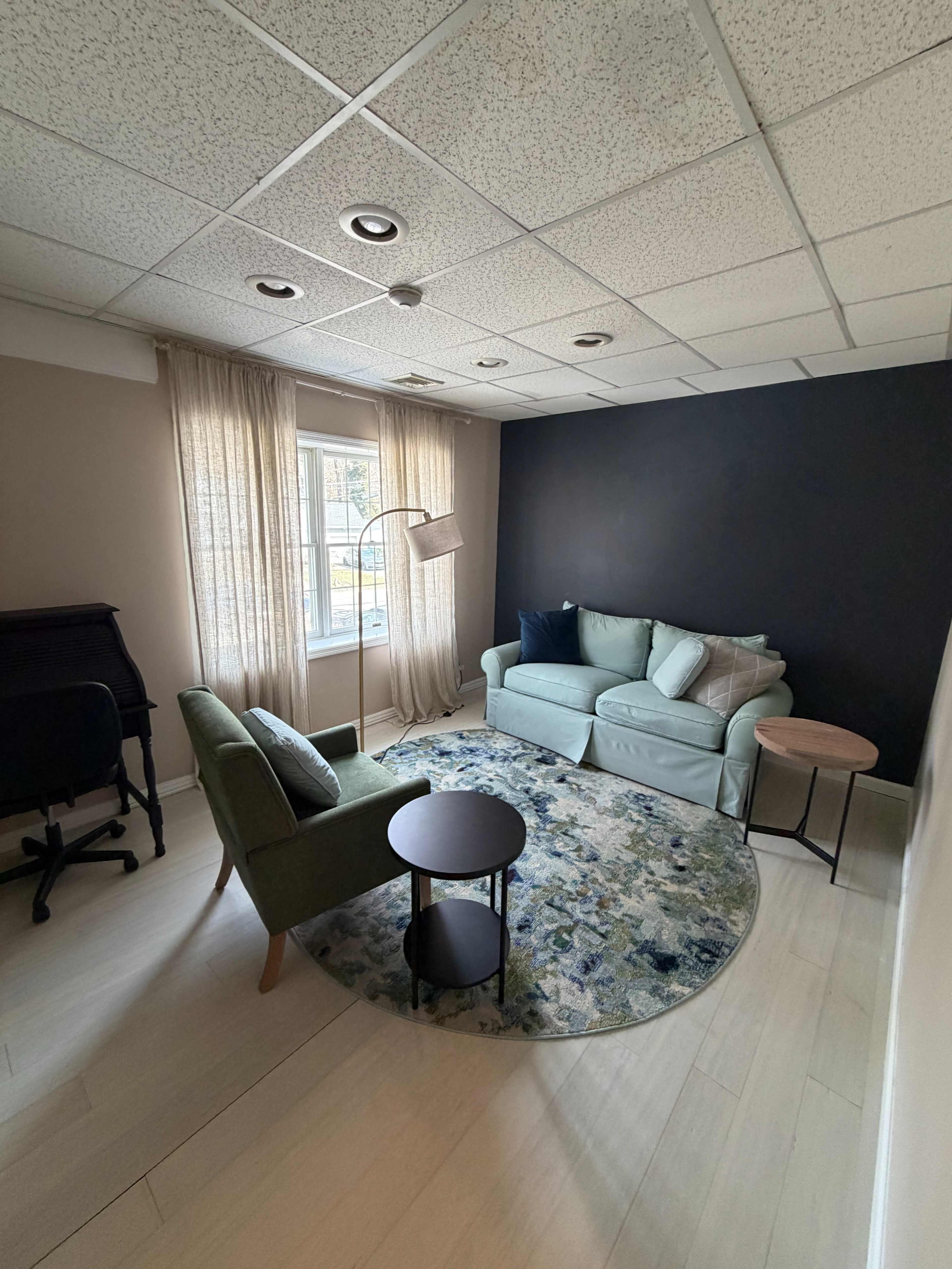The image shows a small lounge area featuring a green couch, two chairs, a round table, and a window draped with light curtains.