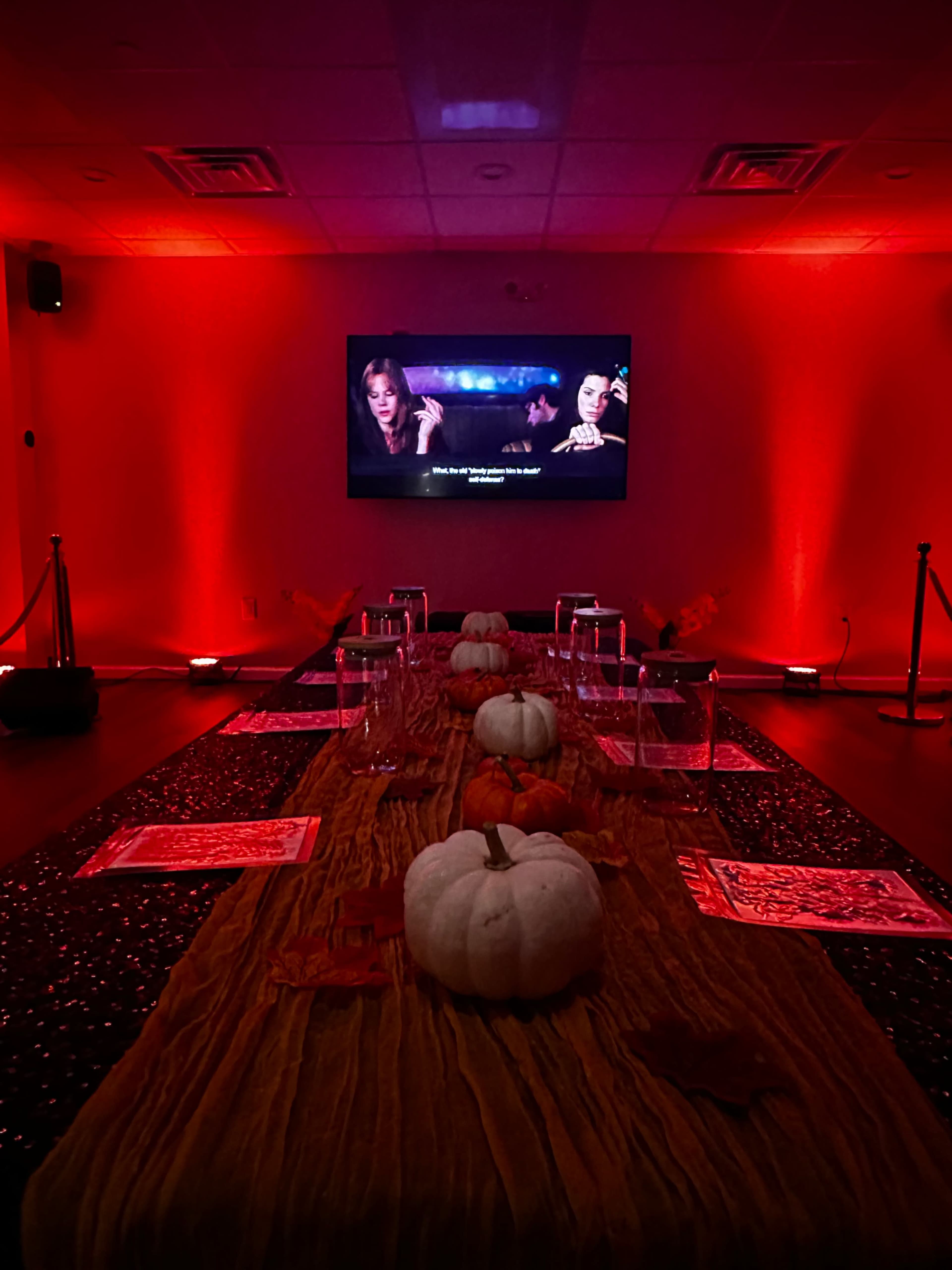 A dimly lit room features a long table decorated with pumpkins and a red backdrop, while a movie plays on a screen.