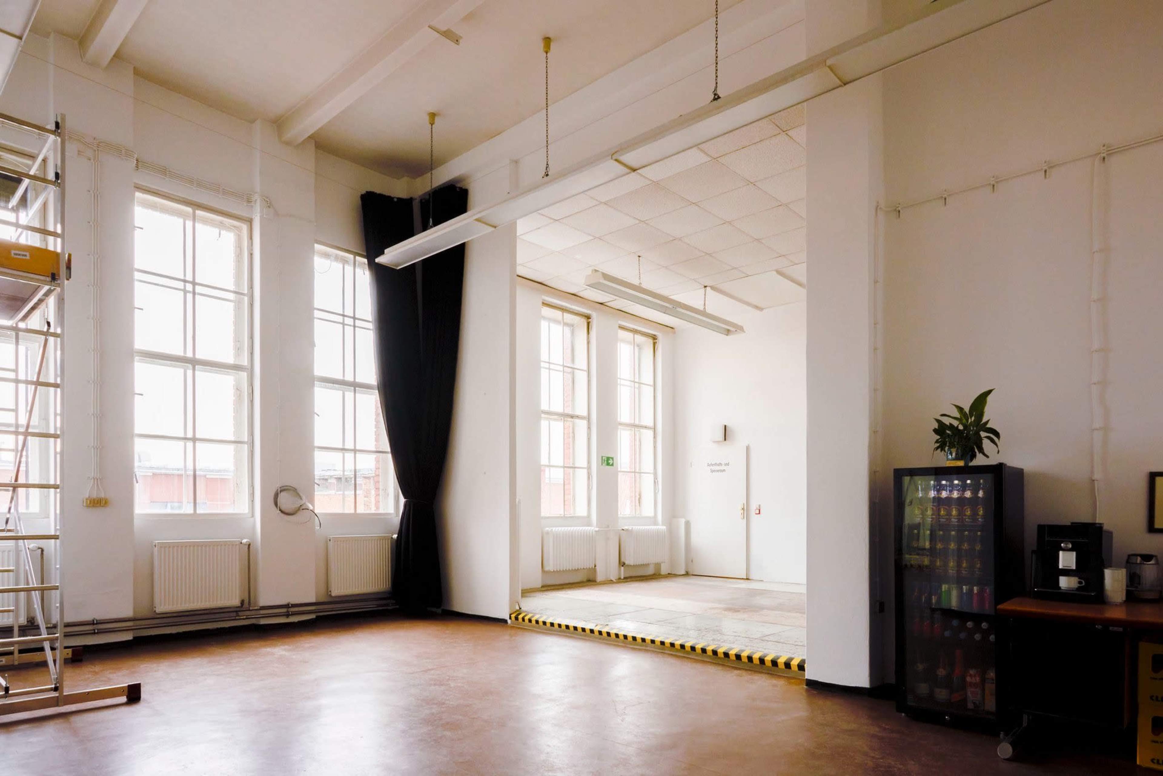 The image shows a spacious, well-lit room with large windows, an empty area at the back, and a kitchenette to the right.