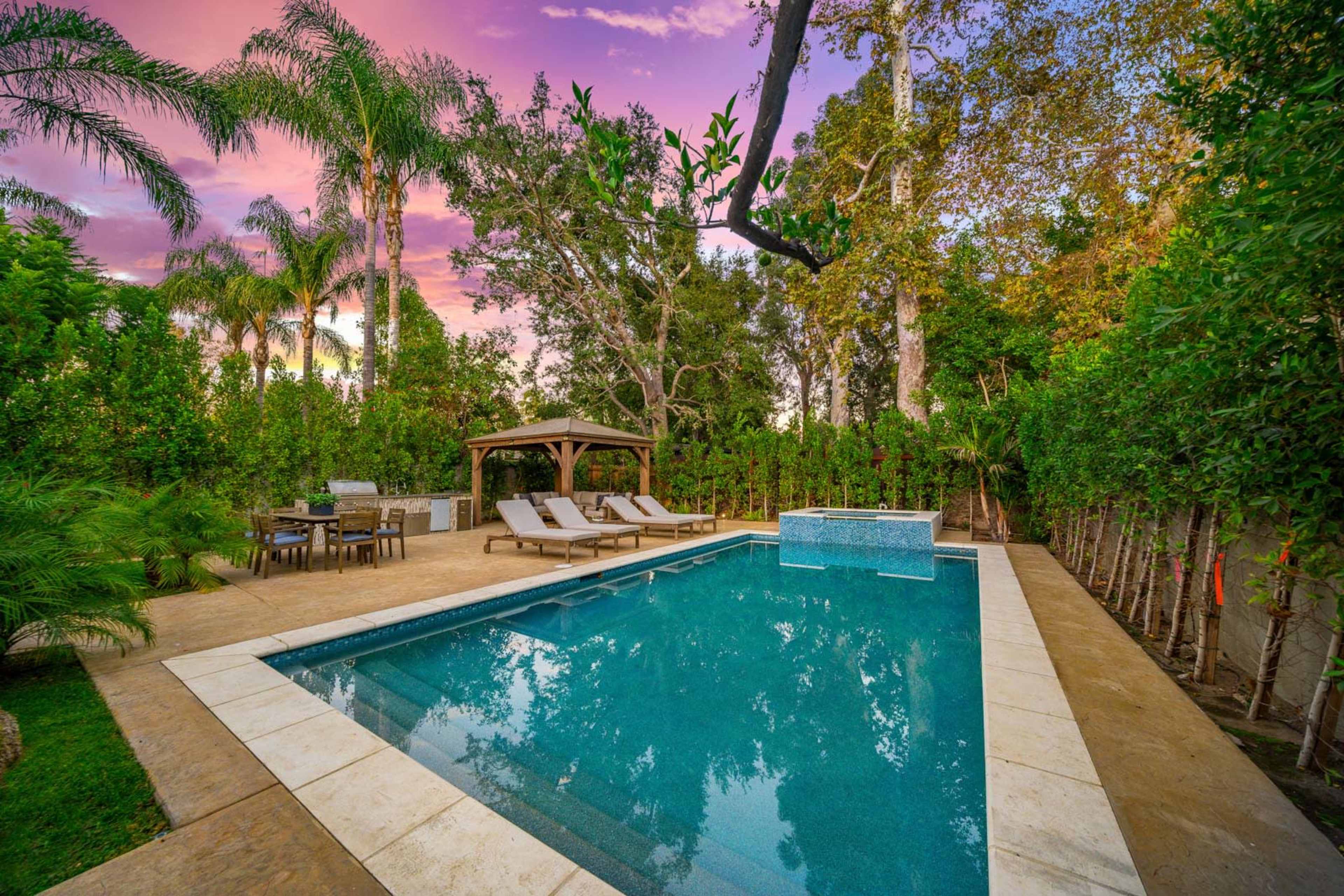The image shows a spacious backyard featuring a swimming pool, lounge chairs, a gazebo, and a dining set surrounded by trees and greenery against a colorful sunset sky.