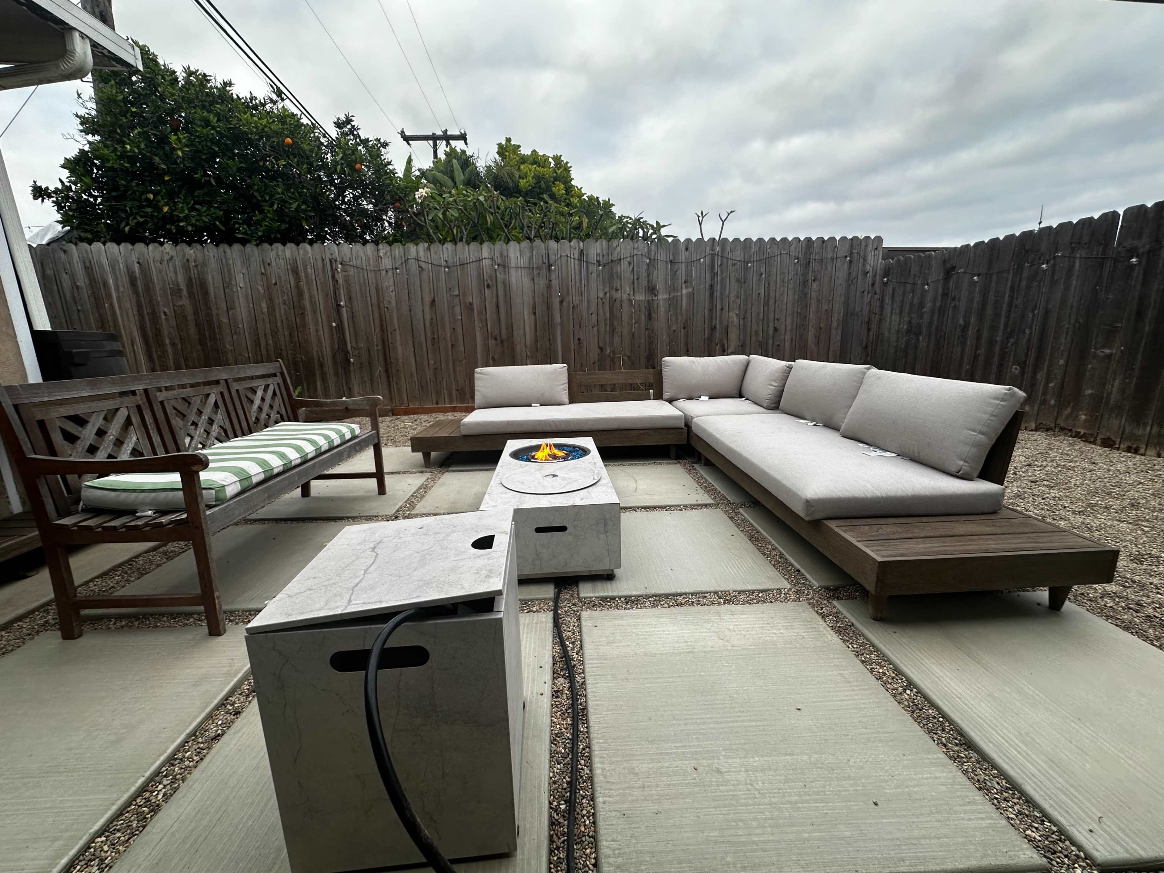 The image shows a patio area with modern outdoor furniture arranged around a fire pit and surrounded by a wooden fence.