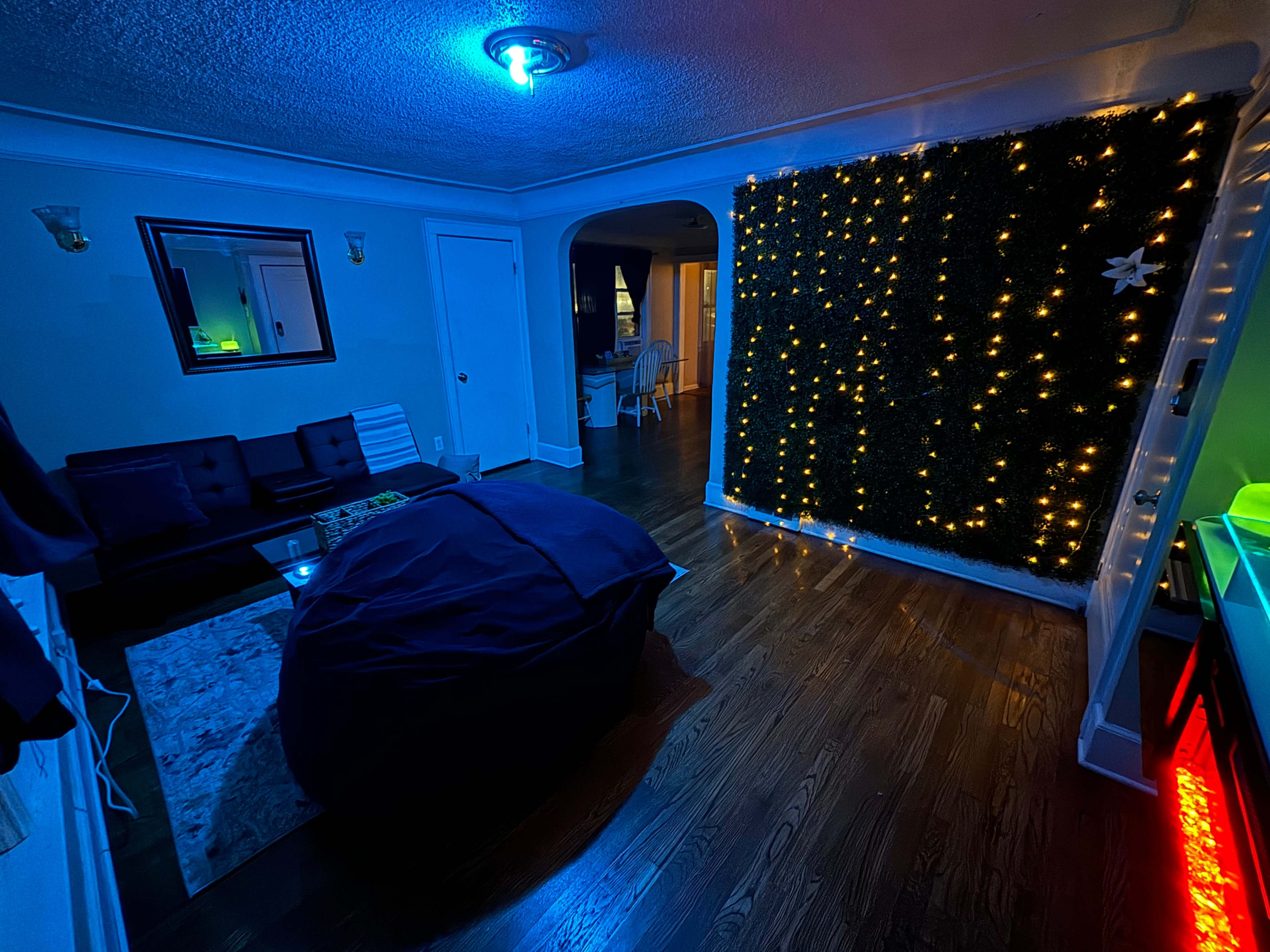 The room features a dark seating area with a bean bag chair and a wall adorned with greenery and string lights, illuminated by cool blue lighting.