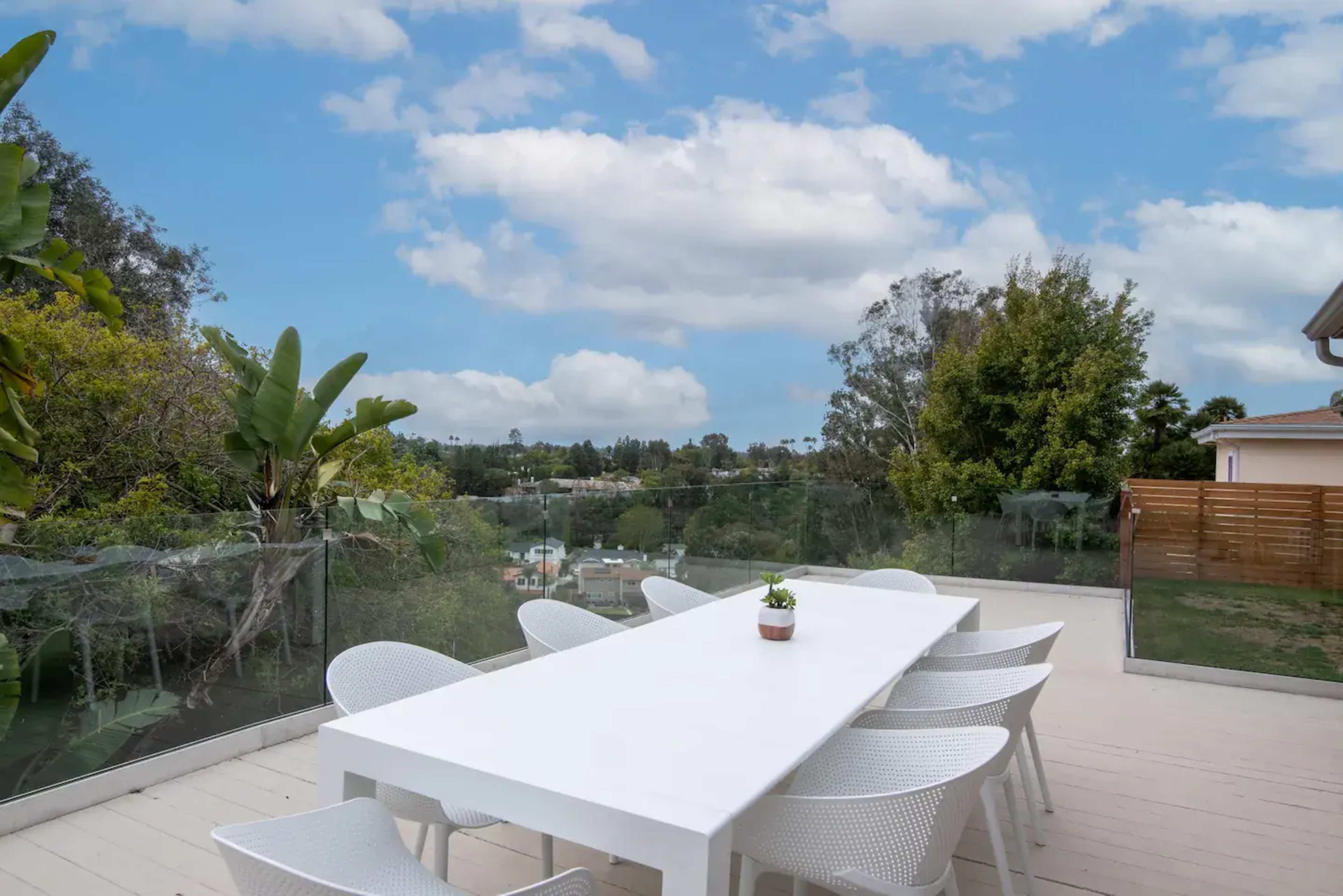 A modern outdoor dining area features a long white table surrounded by chairs, with a view of greenery and a blue sky in the background.
