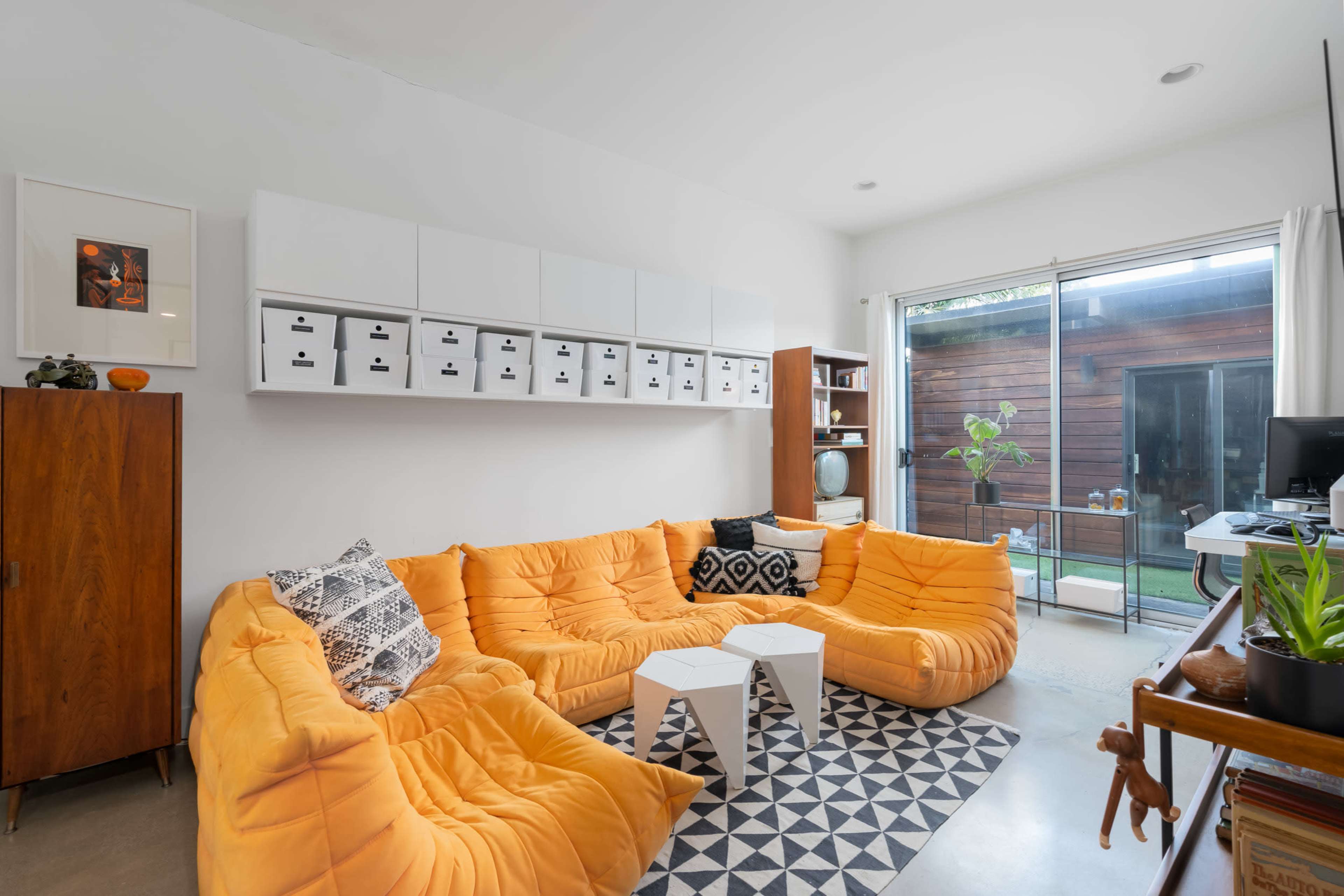 The image shows a cozy living room featuring a large orange sectional sofa, a geometric patterned rug, and white storage boxes mounted on the wall.