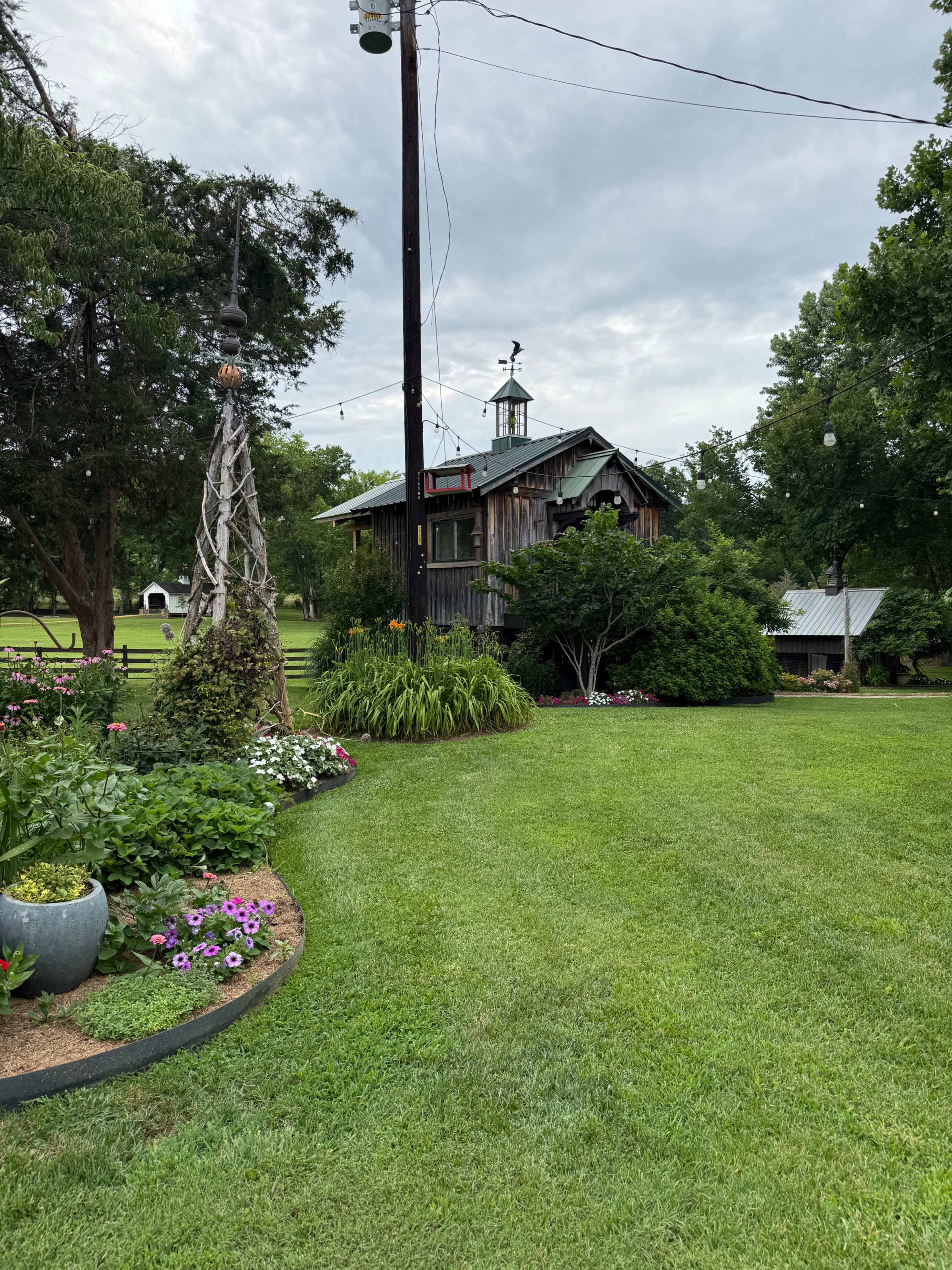 A rustic wooden building with a cupola sits amidst a well-maintained garden featuring flowers and manicured lawns, under a cloudy sky.