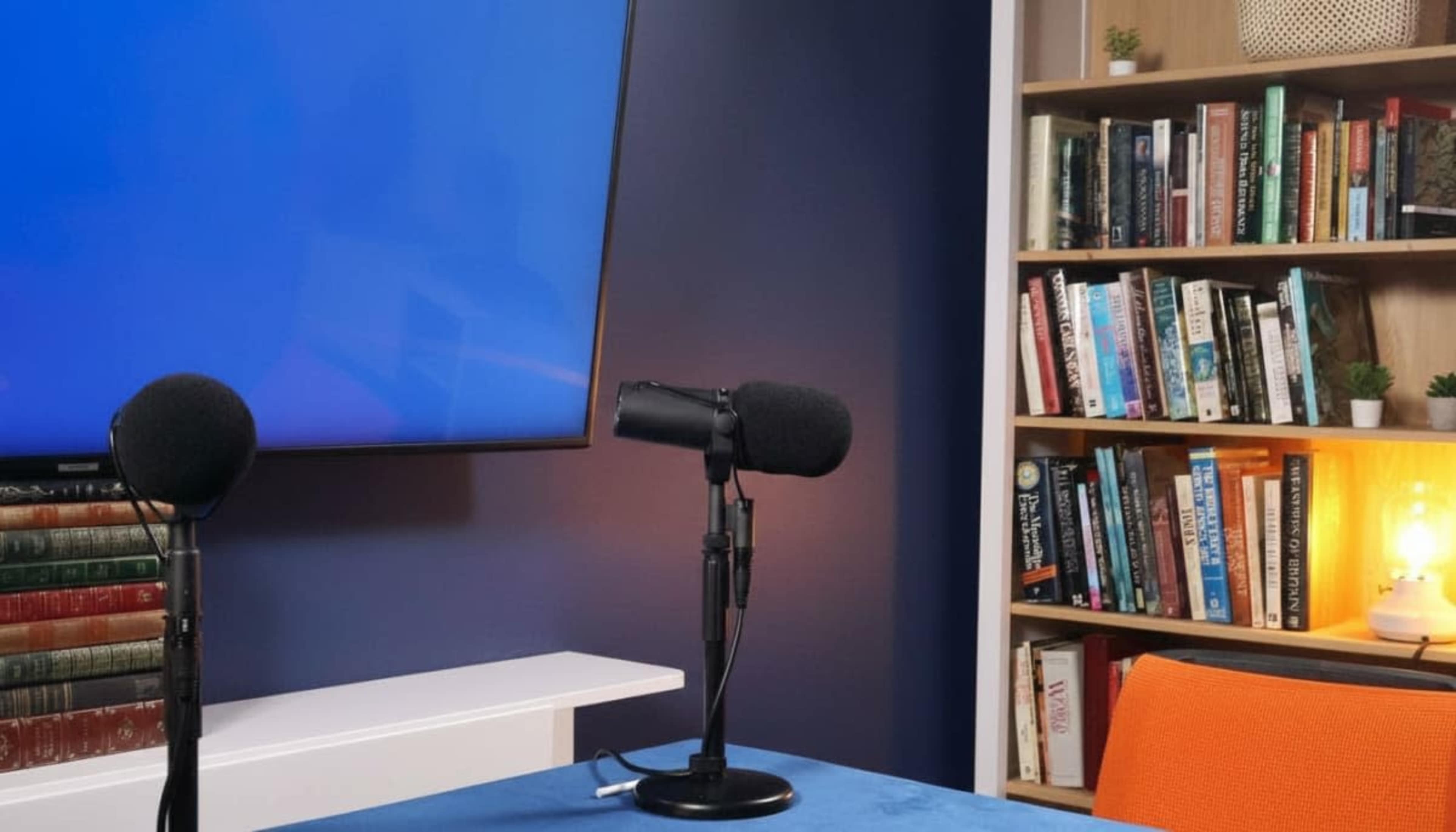 The image shows a podcasting setup with two microphones on stands, a large screen displaying a blue background, and a bookshelf filled with books illuminated by a warm light.