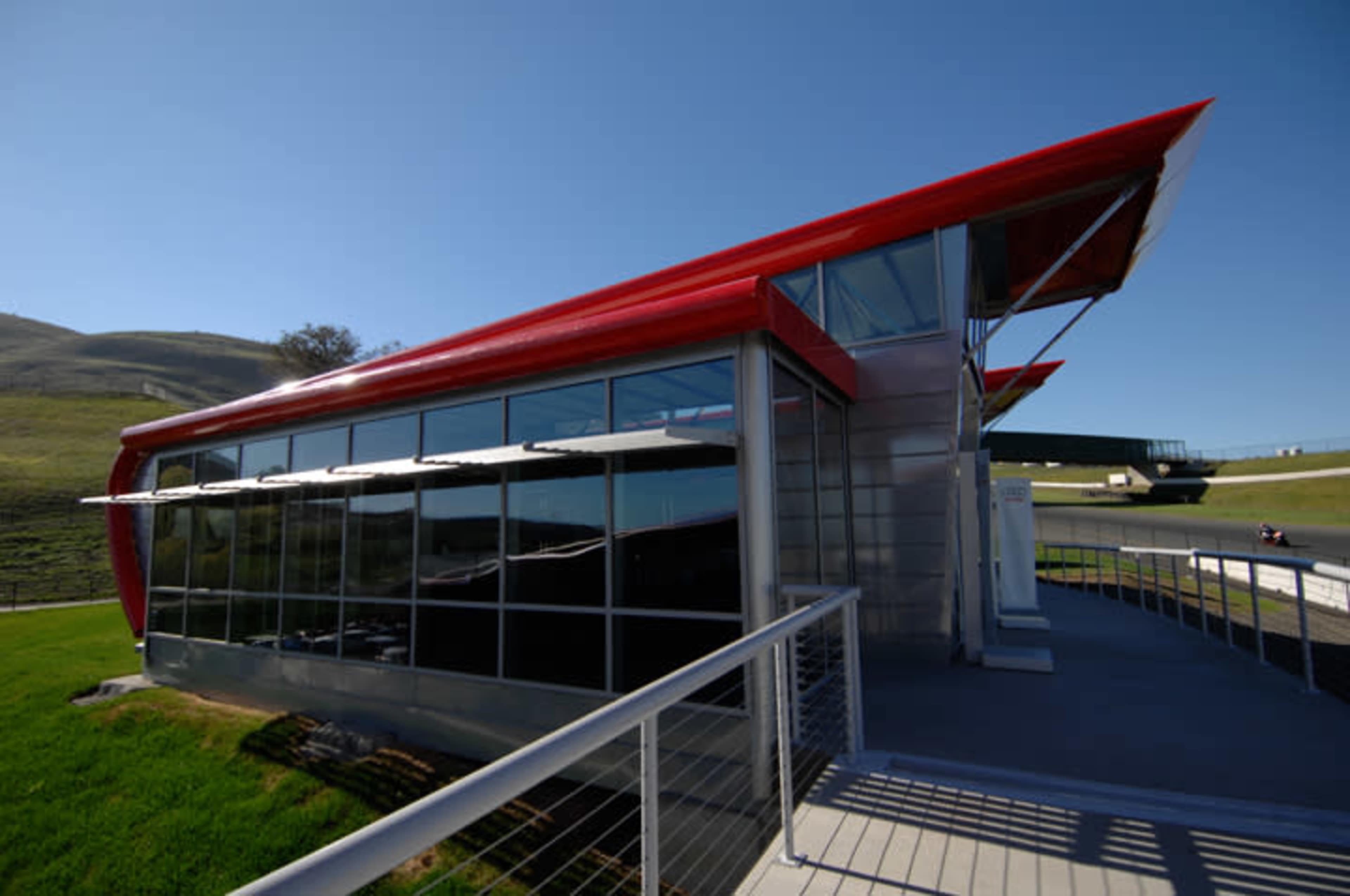A modern building with a distinctive red roof and large glass windows, located near a racetrack surrounded by green hills.