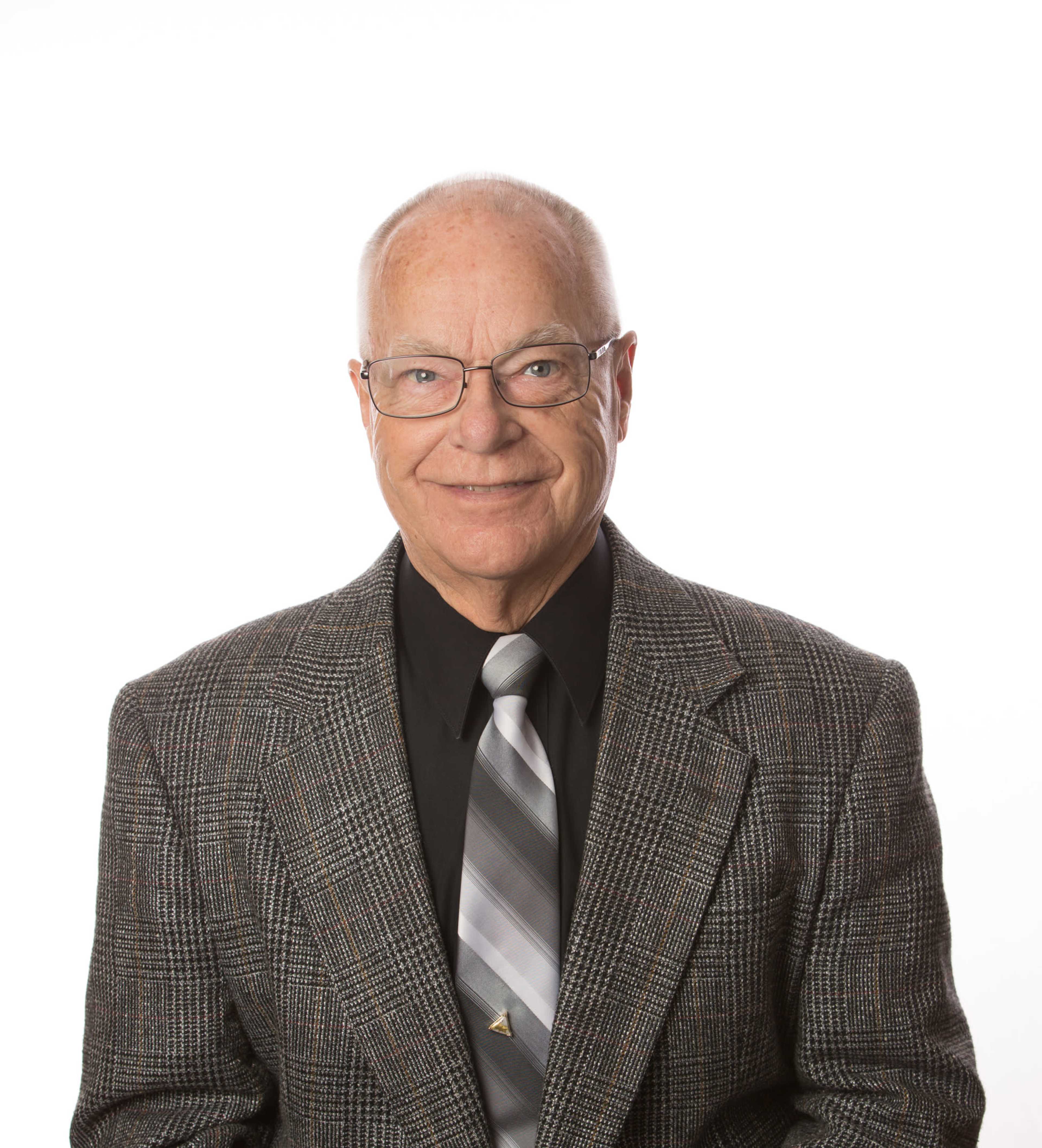 The image shows an older man wearing glasses and a suit with a patterned blazer and a tie, smiling against a plain white background.