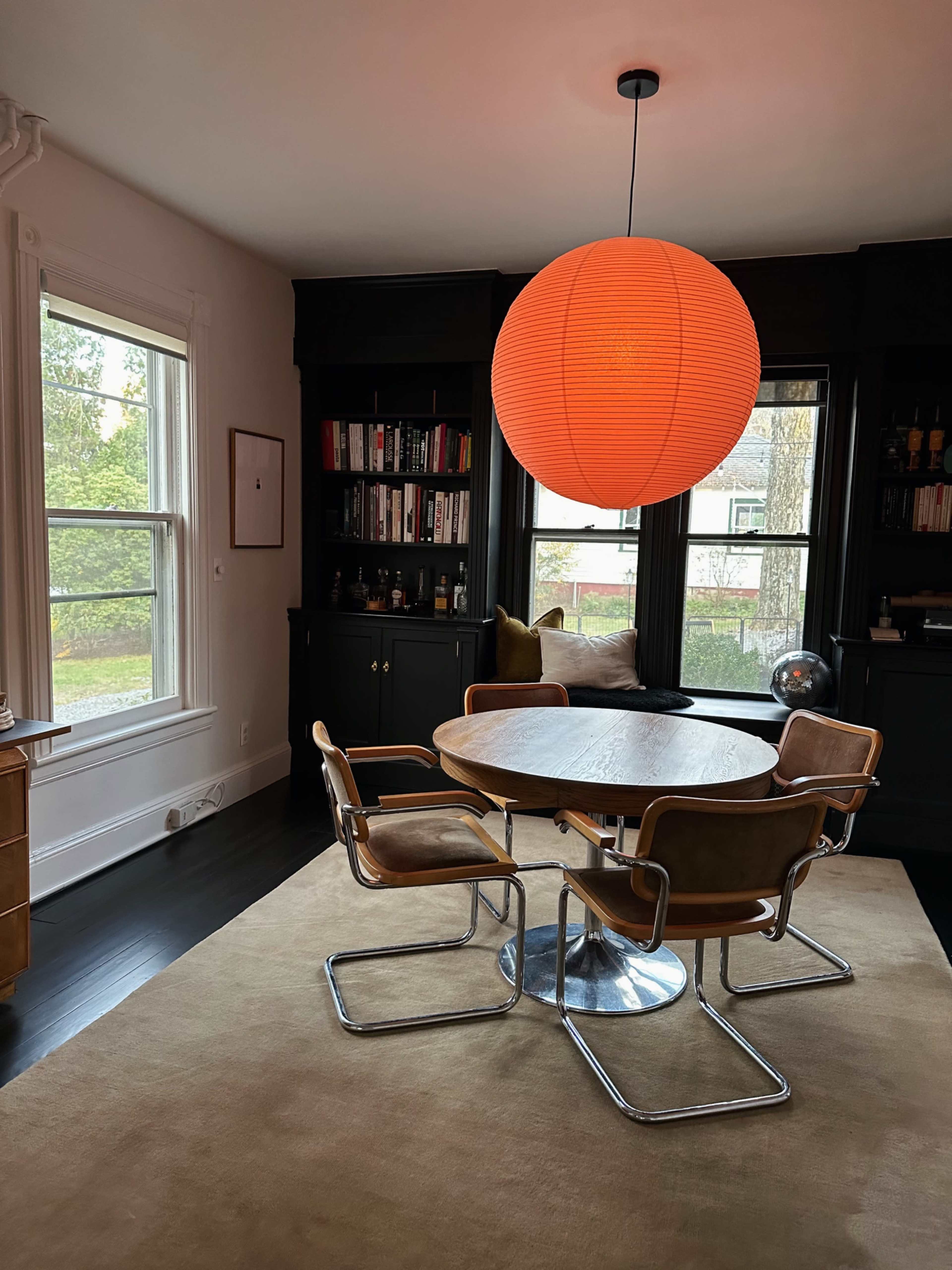 A modern dining area featuring a round wooden table surrounded by four leather chairs beneath a large orange pendant light, with a bookshelf on one side and windows letting in natural light.