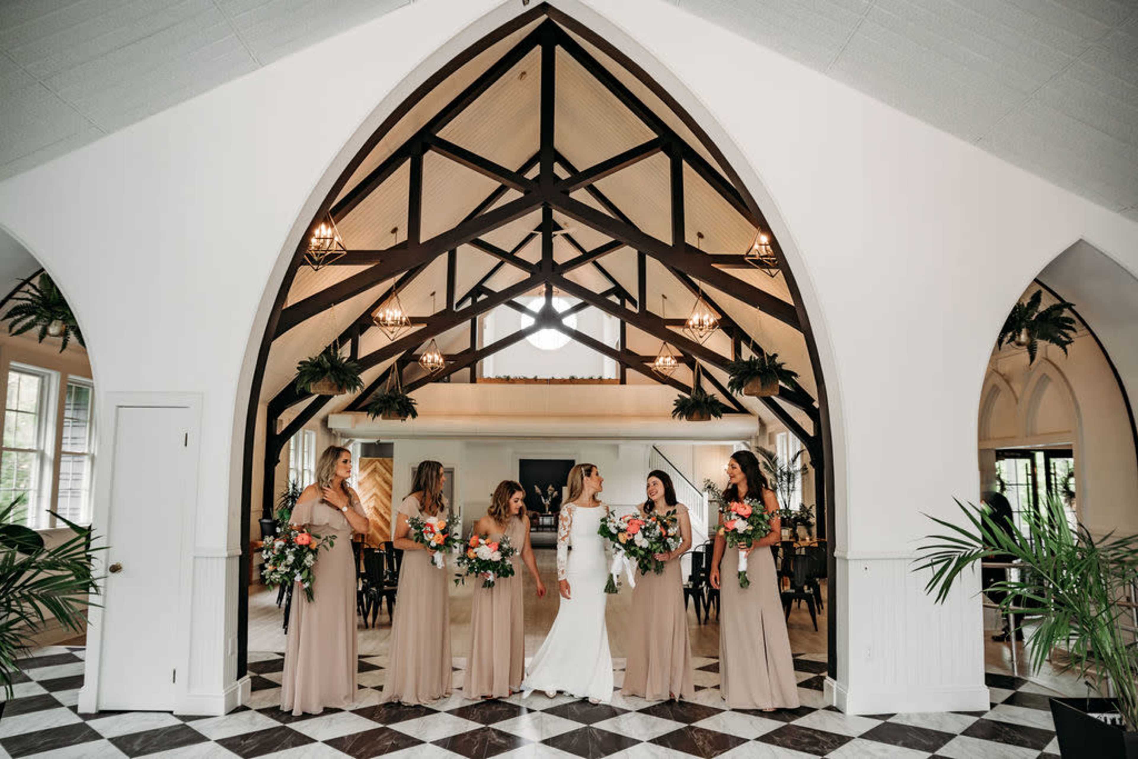 A bride and her bridesmaids stand together in a spacious hall featuring an arched ceiling and patterned tile flooring.