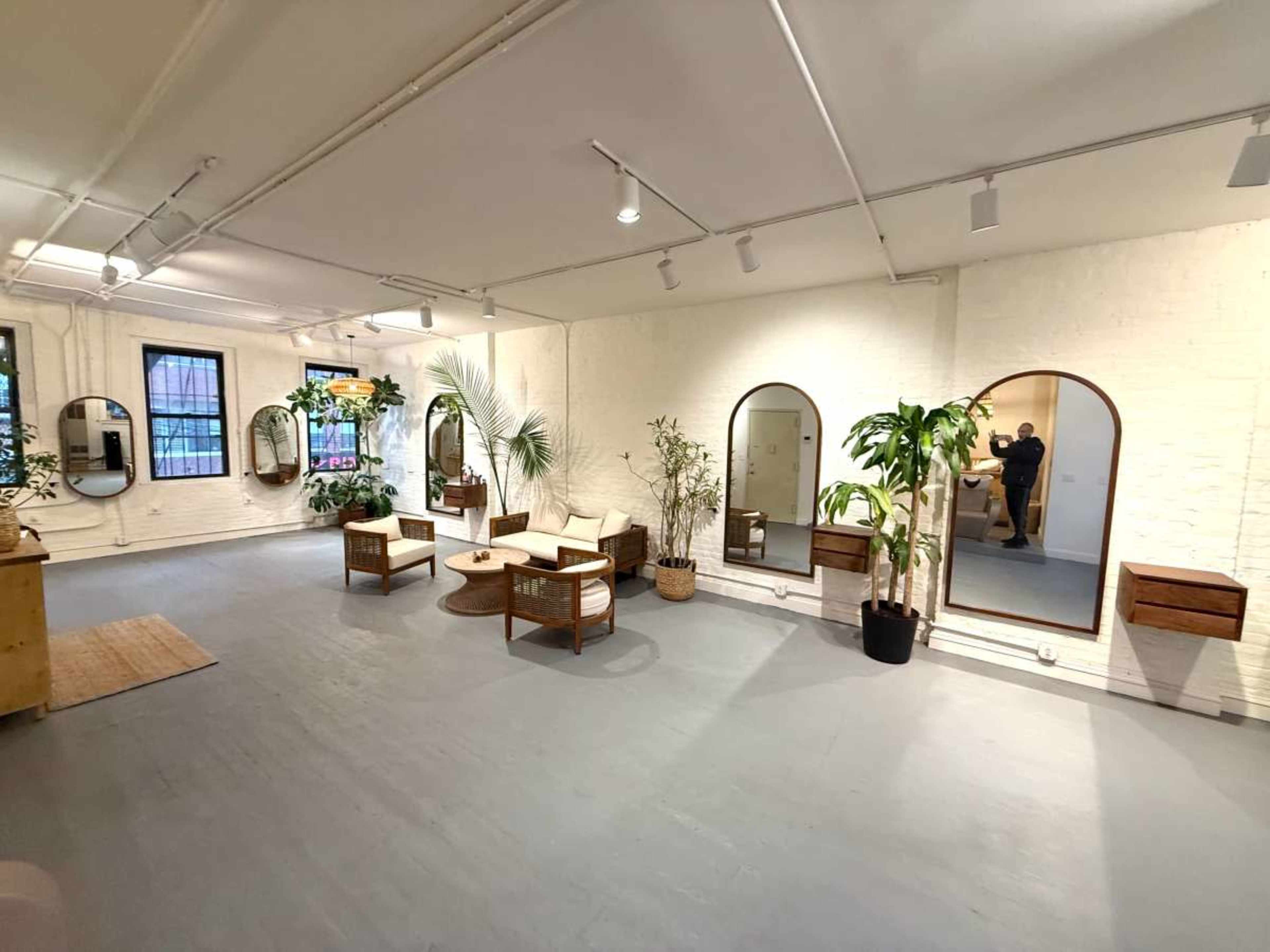 The image shows a spacious room with white walls, multiple mirrors, green plants, and a seating area featuring chairs and a table.
