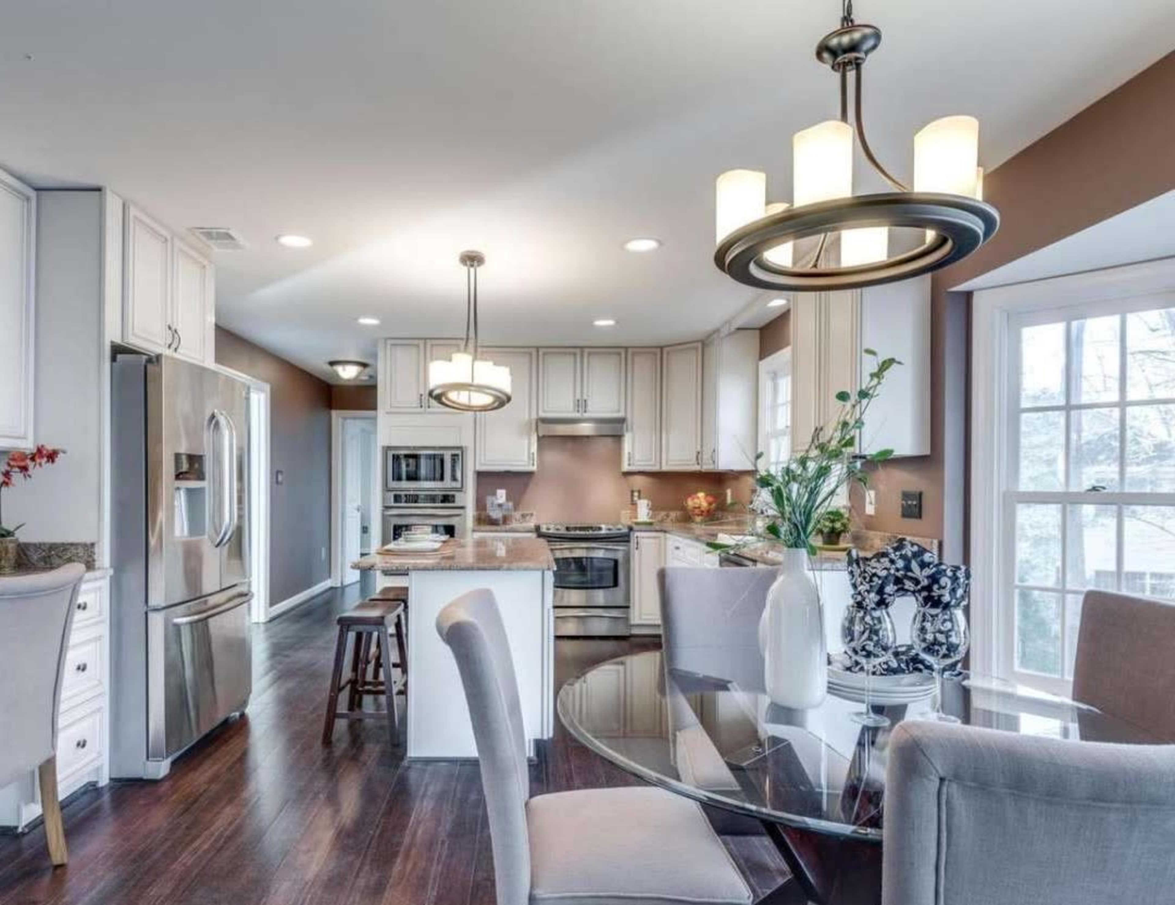 The image shows a modern kitchen and dining area with light-colored cabinets, stainless steel appliances, and a glass dining table surrounded by upholstered chairs.