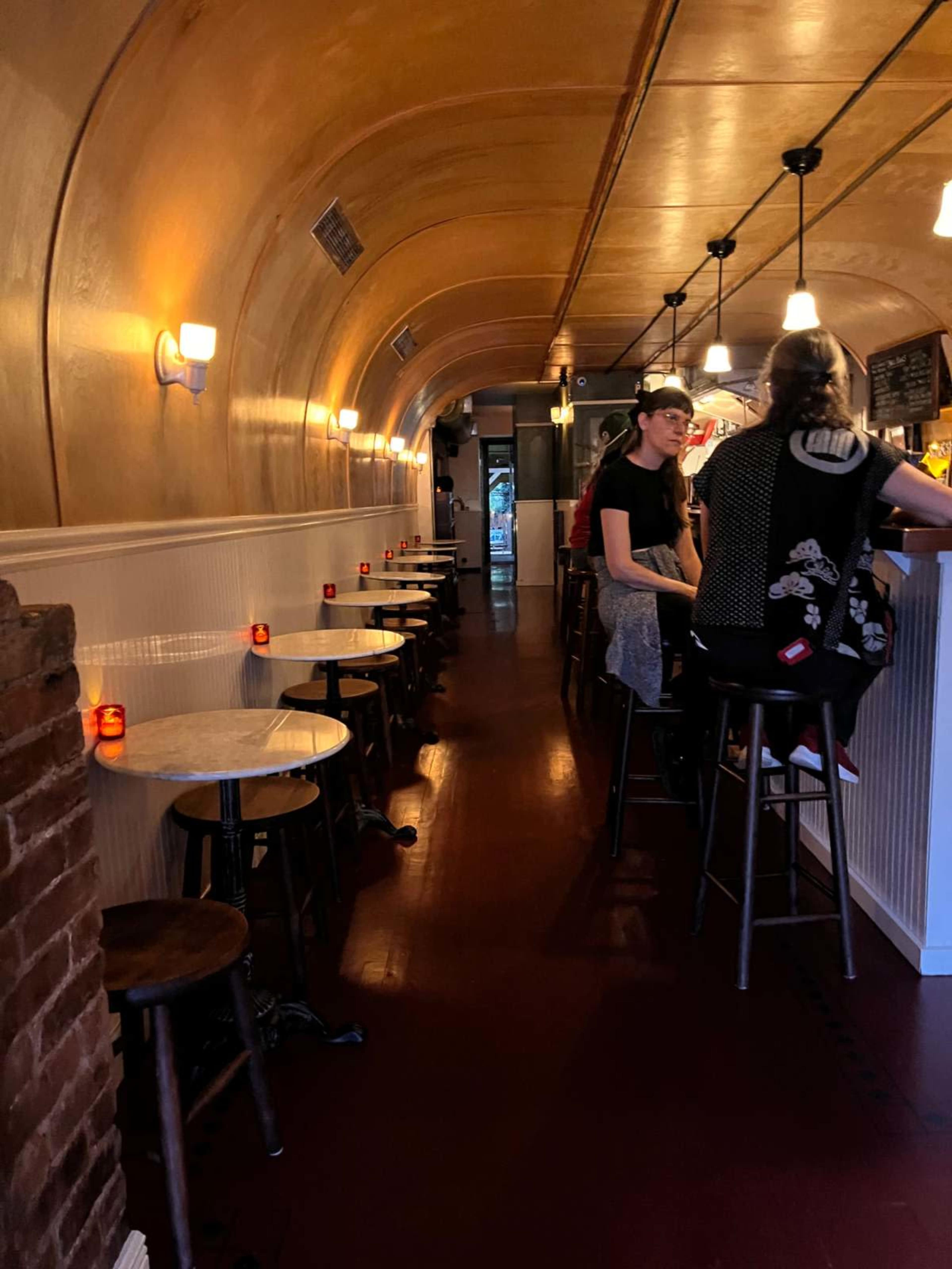 The image shows a cozy bar with round tables and stools lining a narrow, illuminated corridor, where patrons are seated and a bartender serves drinks.
