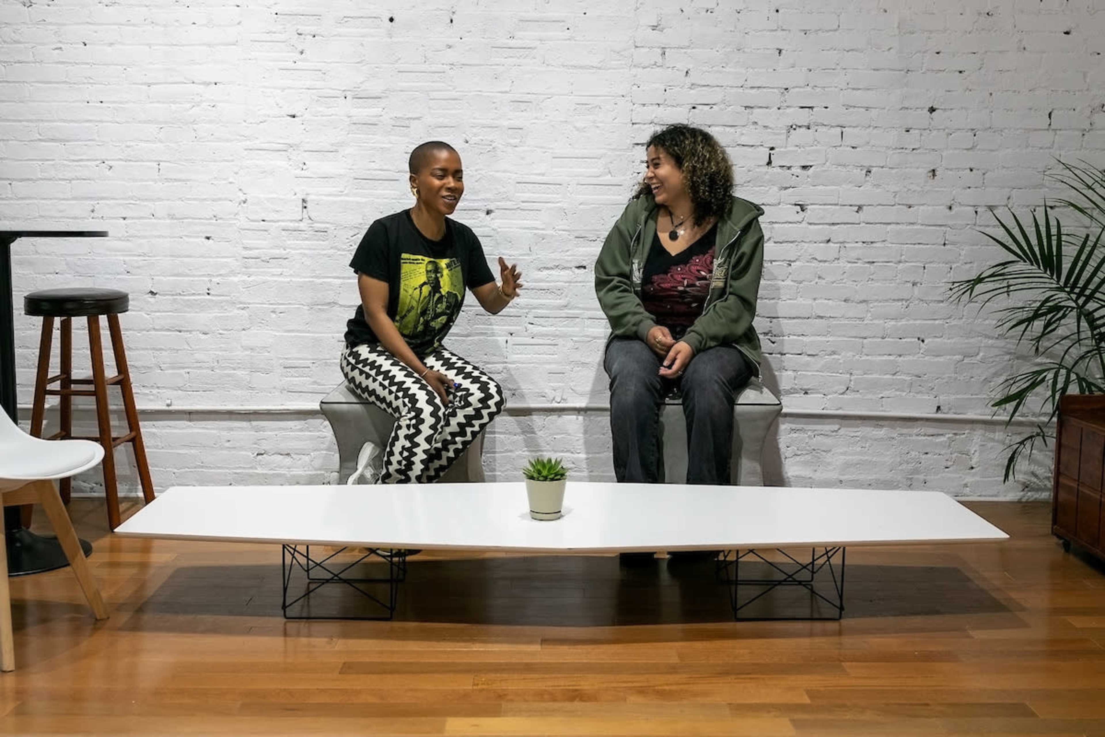 Two women are sitting on a bench in a minimalist room with a white brick wall and a small plant on a long table between them.