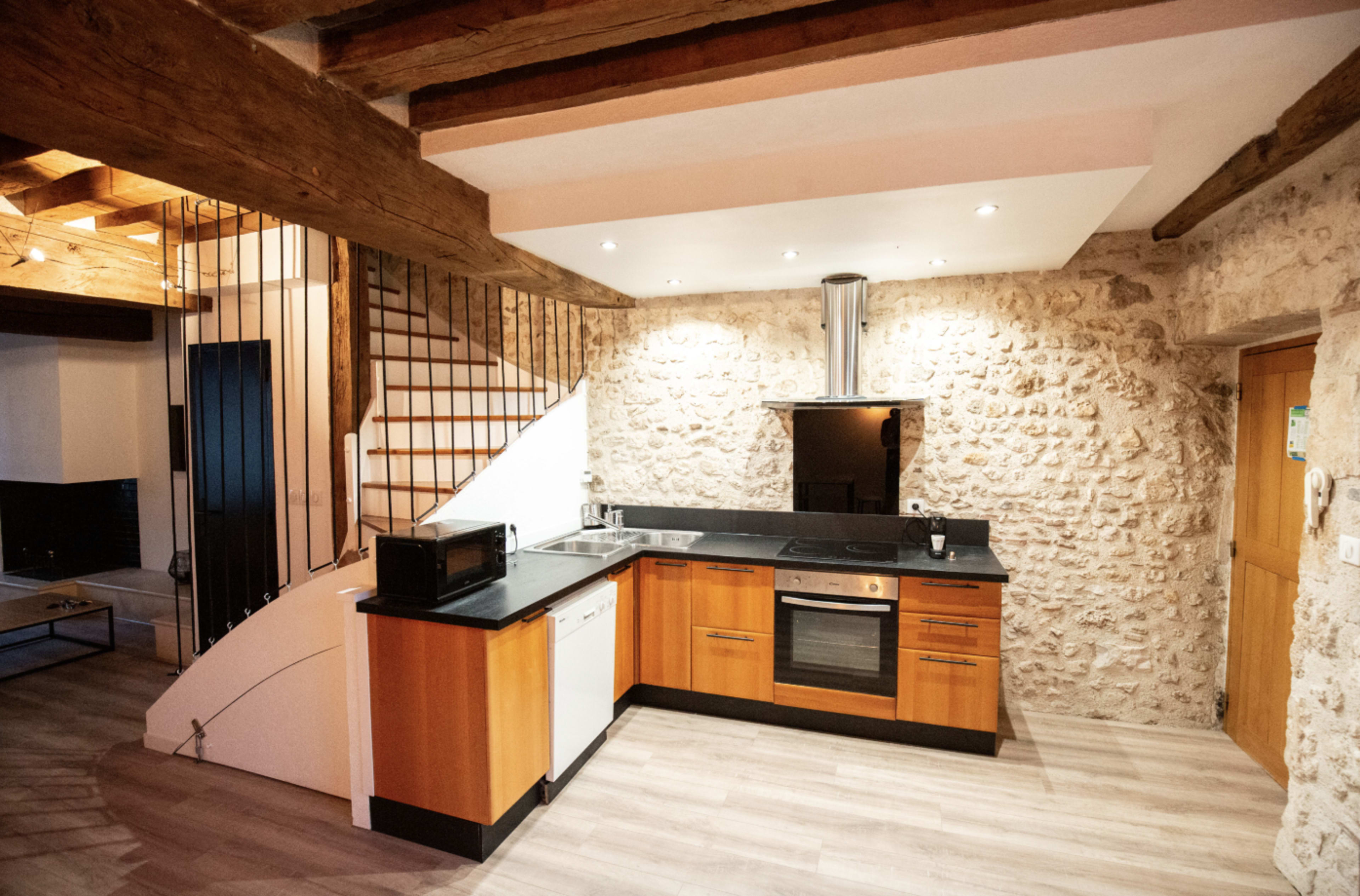 The image shows a modern kitchen featuring a black countertop, wooden cabinets, and exposed stone walls, with a staircase visible in the background.