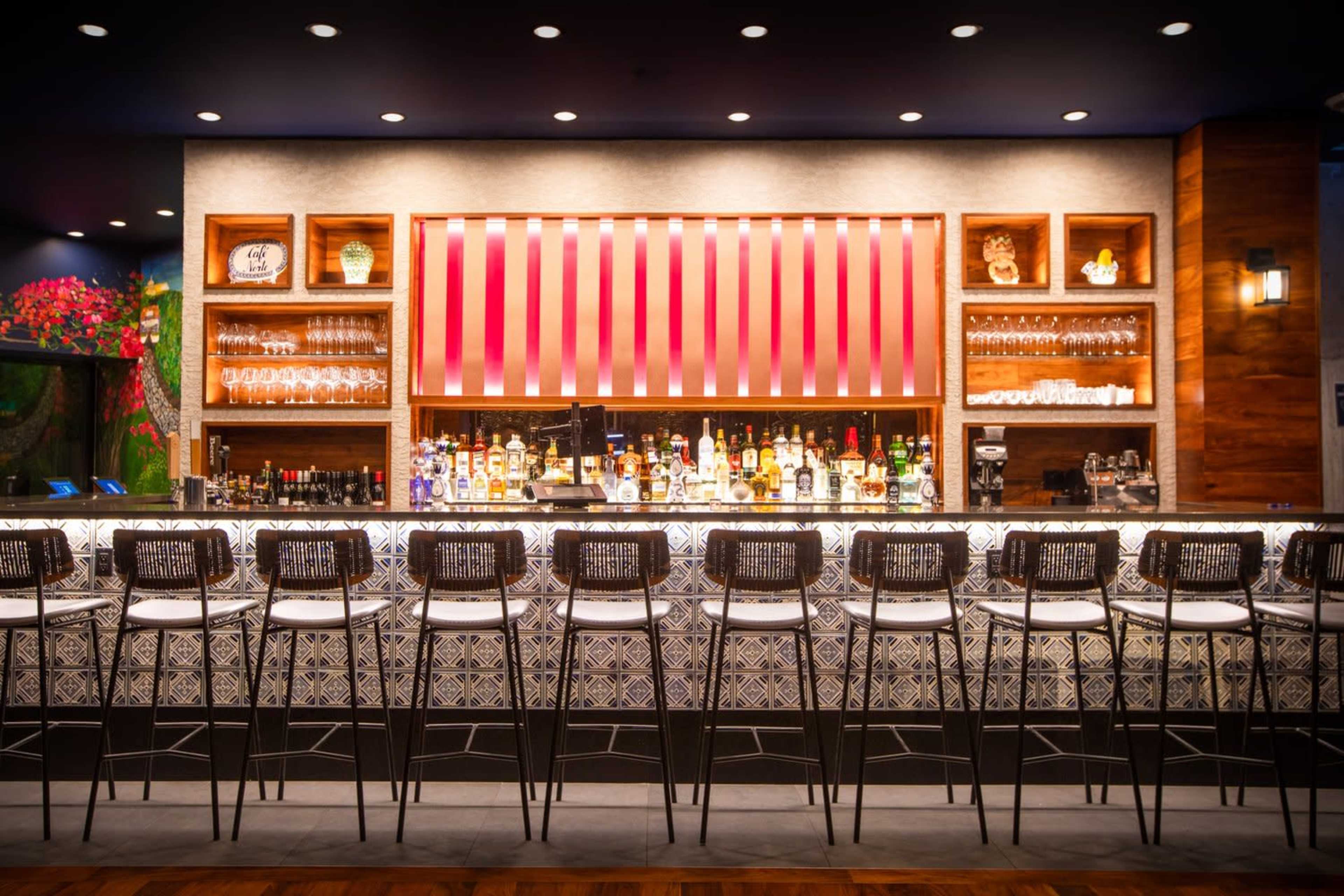 A modern bar features a long counter with high-backed stools in front of a well-stocked display of colorful liquor bottles and glassware against a stylish backdrop.