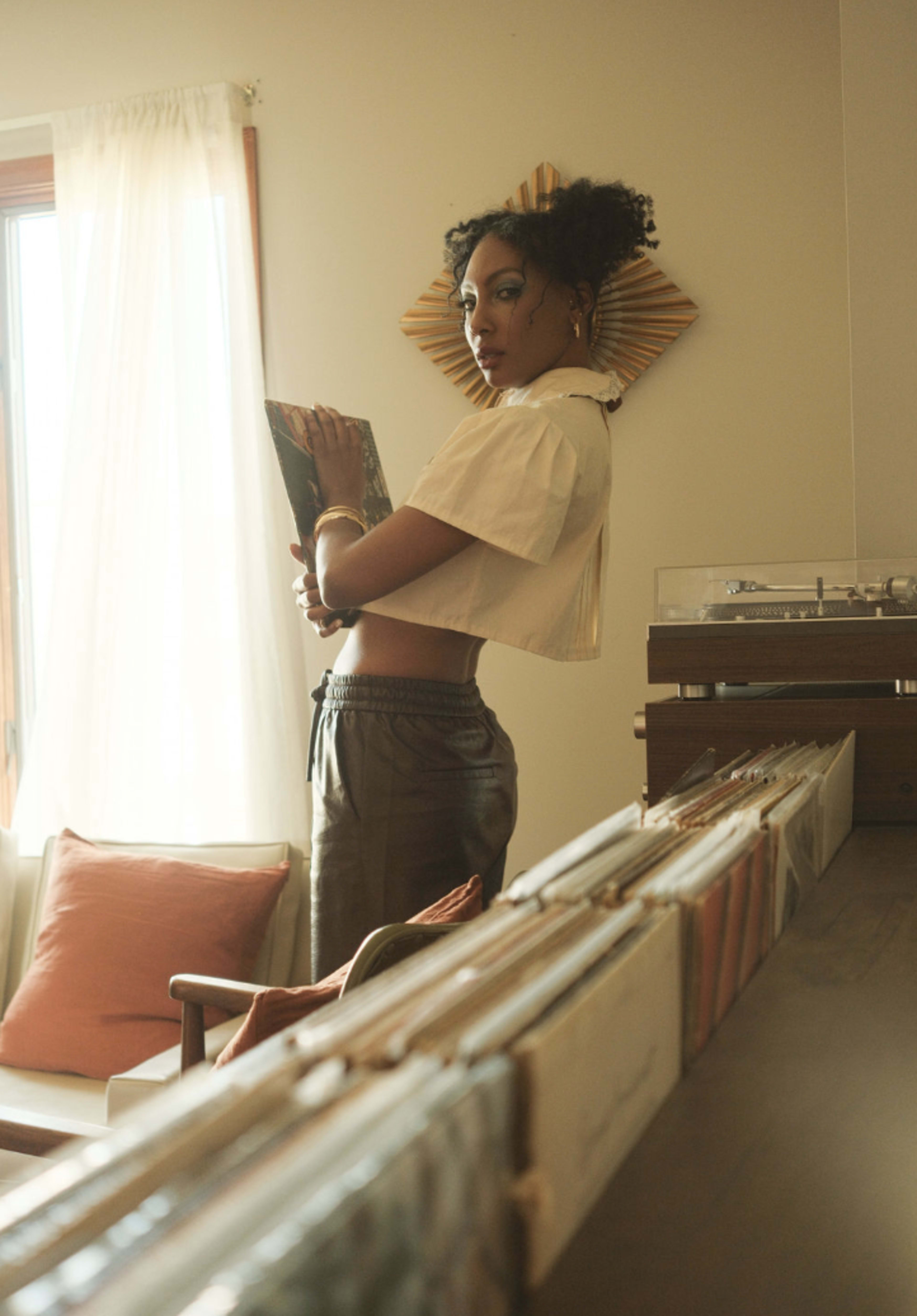 A person with curly hair stands in a room holding a record album, with a row of vinyl records visible in the foreground.