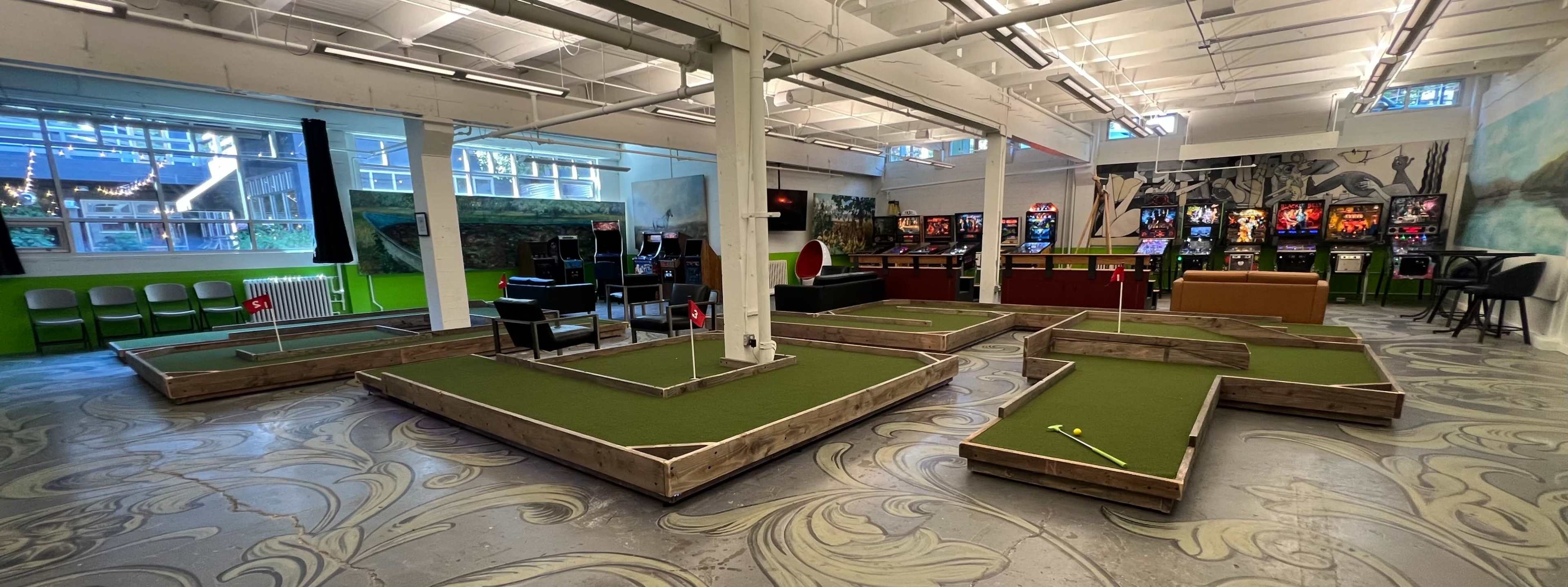 The image shows an indoor mini-golf venue with several artificial turf putting greens, surrounded by pinball machines and seating areas.