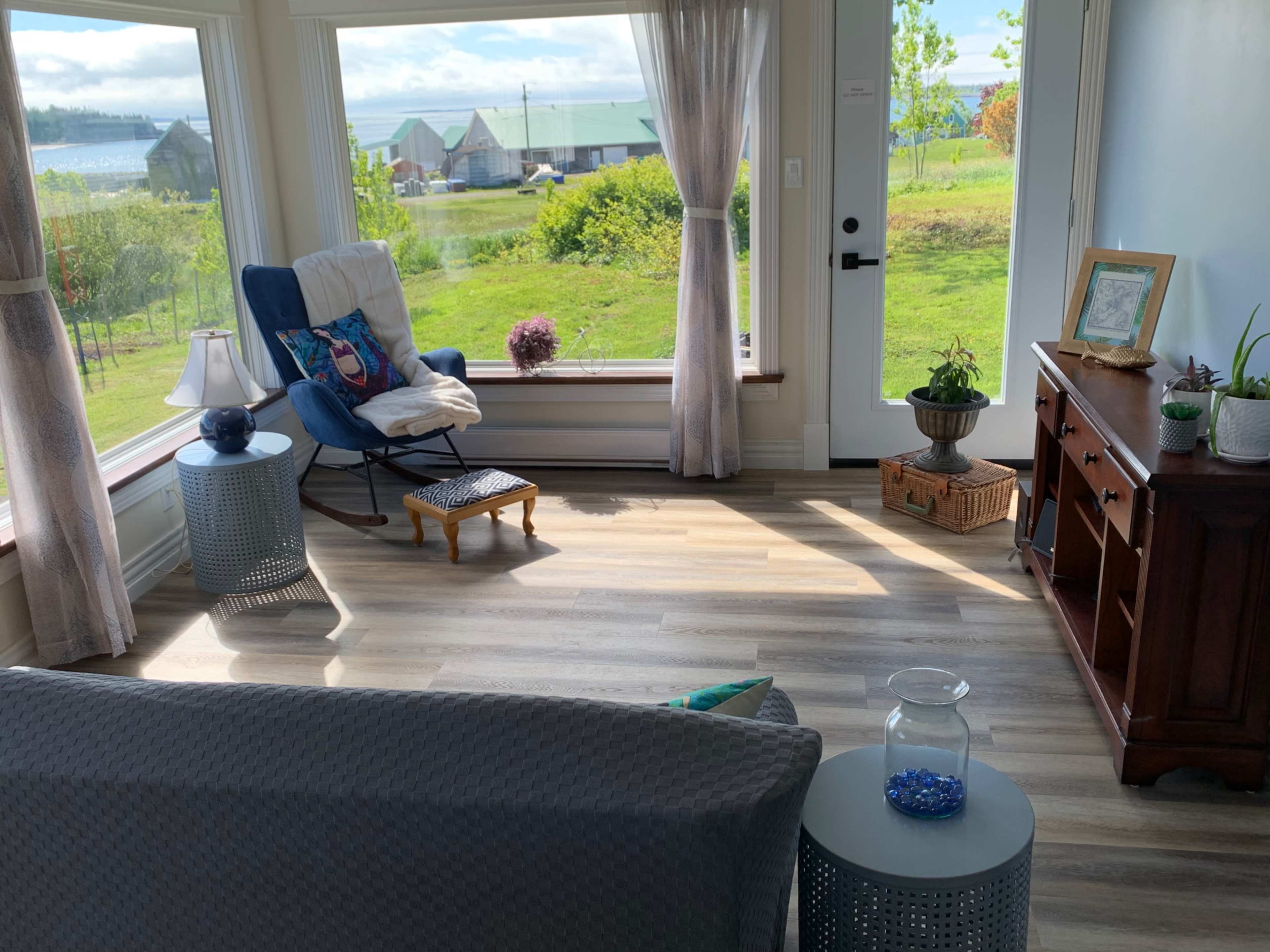 A bright room features large windows with a view of green fields, a blue armchair, a wooden side table, and a console with decorative items.