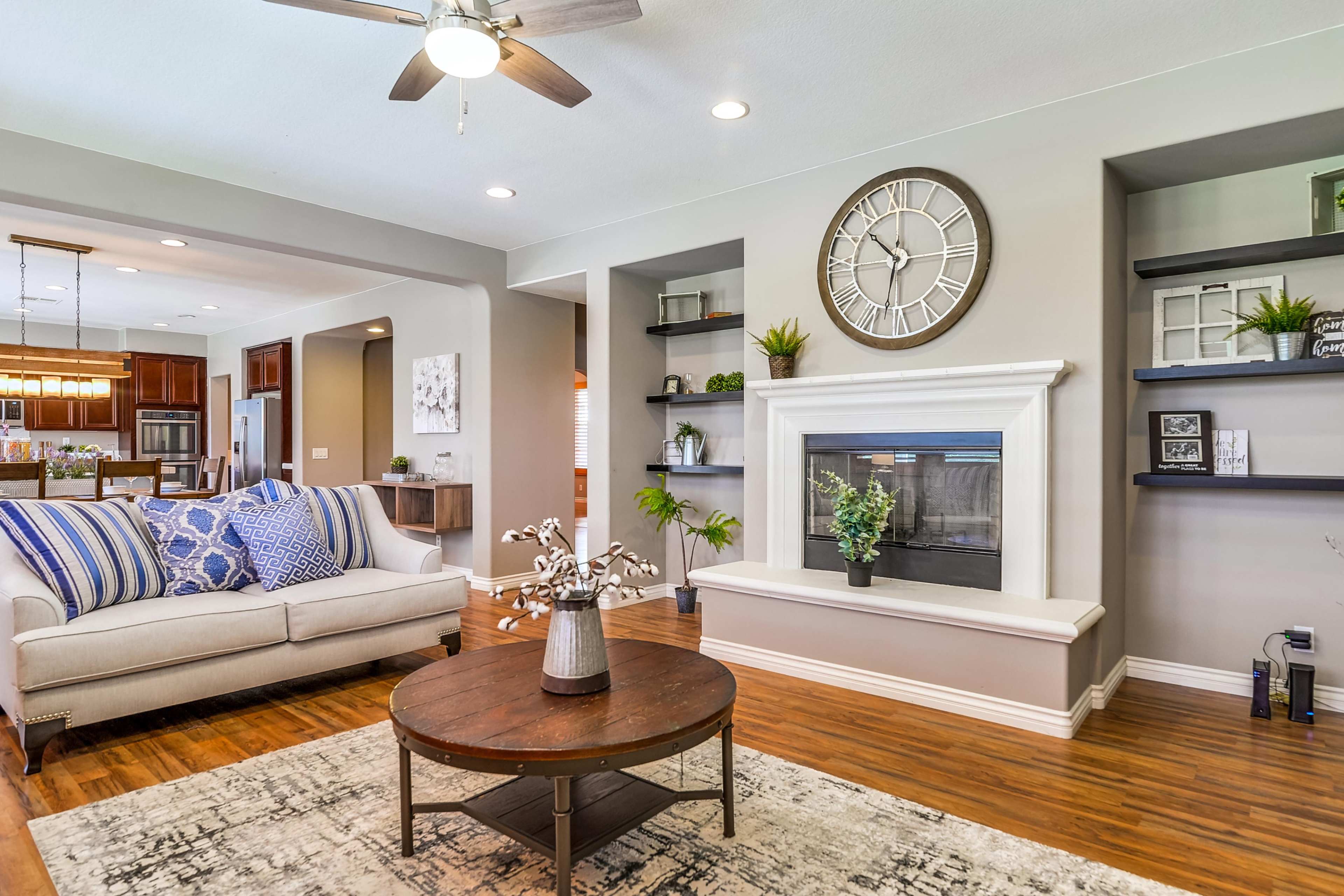 A living room features a white fireplace, a grey wall clock, a round wooden coffee table, and a beige sofa with blue striped throw pillows.