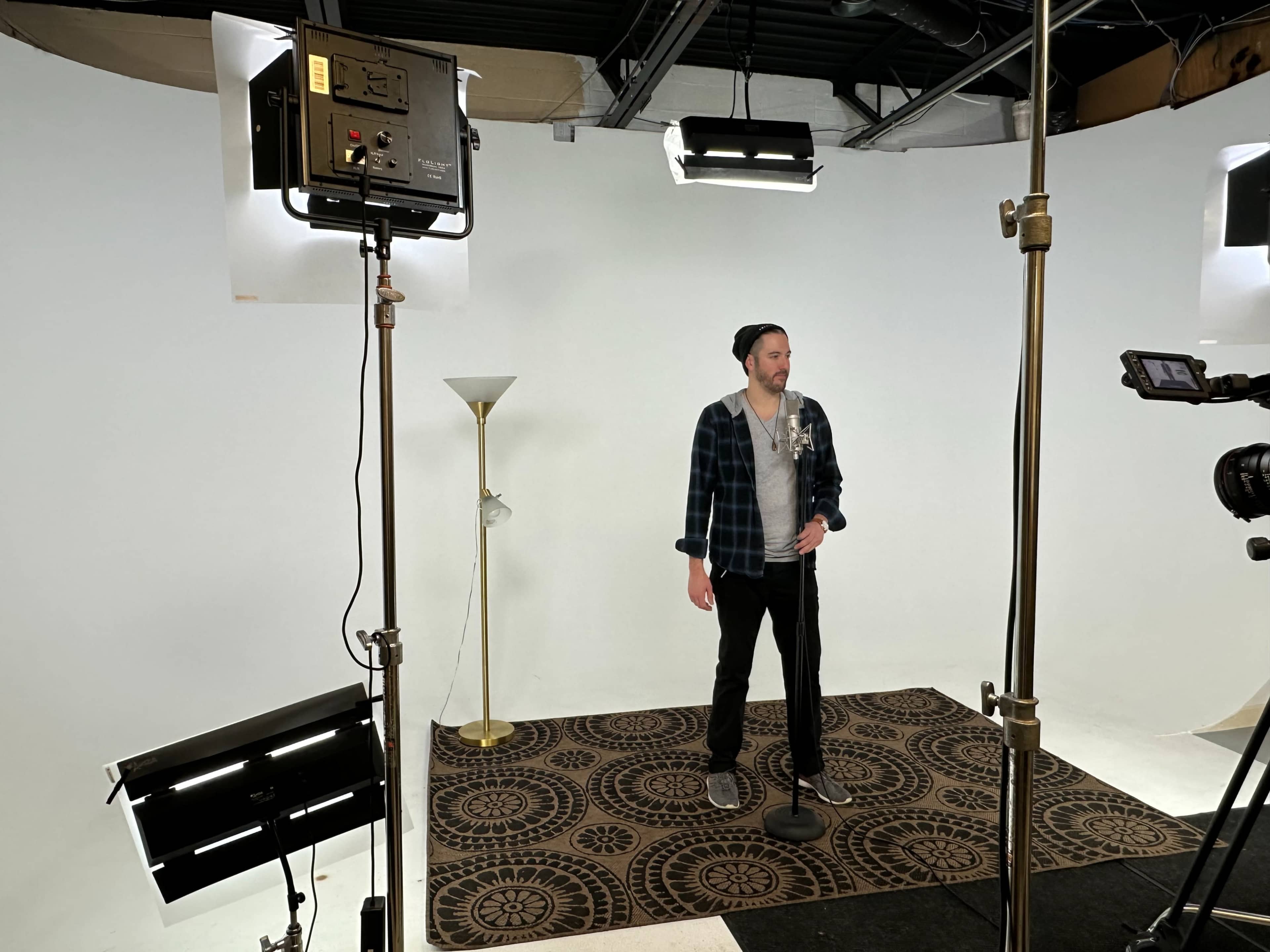 A person stands on a patterned rug in front of a white backdrop, holding a microphone, with studio lights and a camera focused on them.